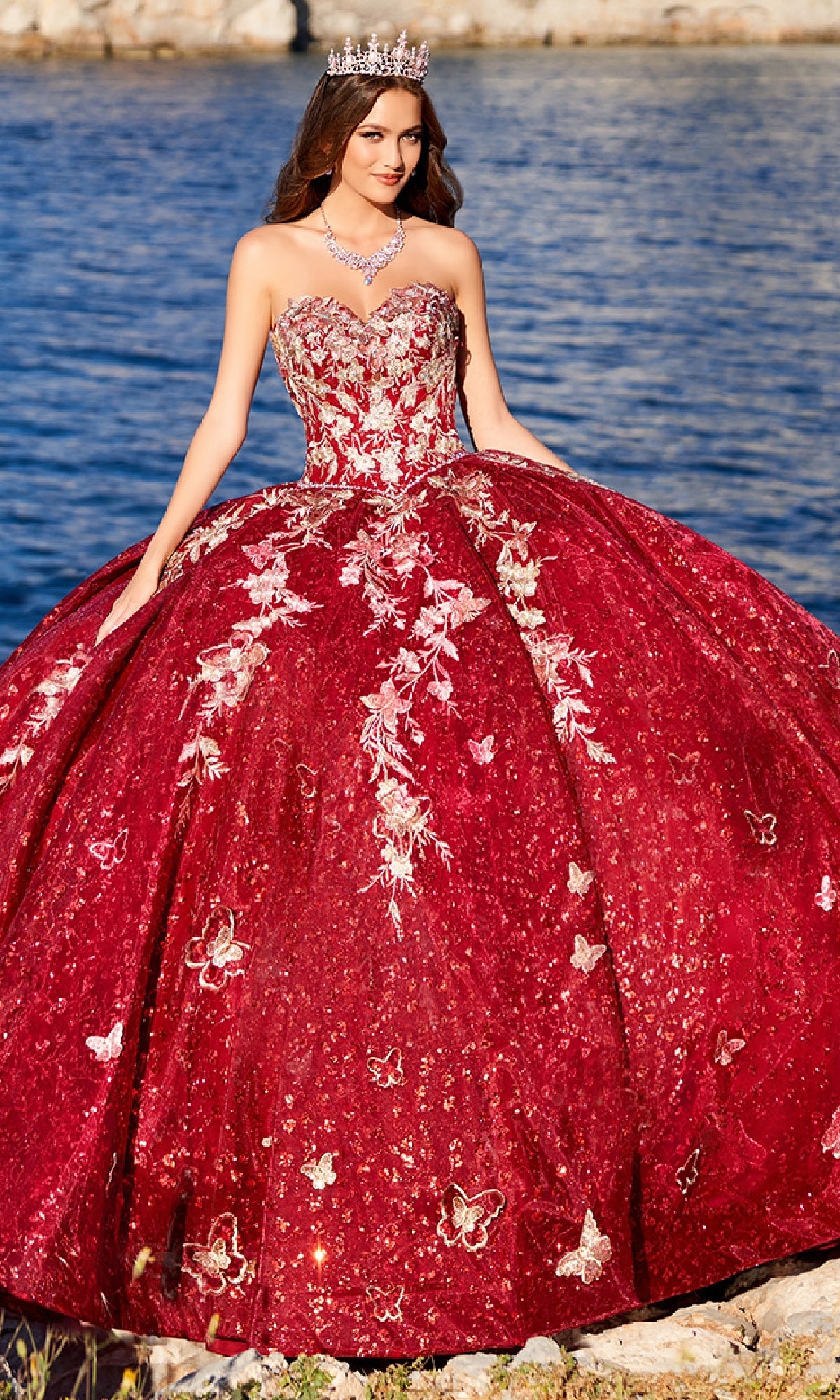 A woman in the strapless Princesa PR30121 Quinceañera dress with gold embroidered lace and crown stands by the water, her sparkling red tulle ball gown catching the light.
