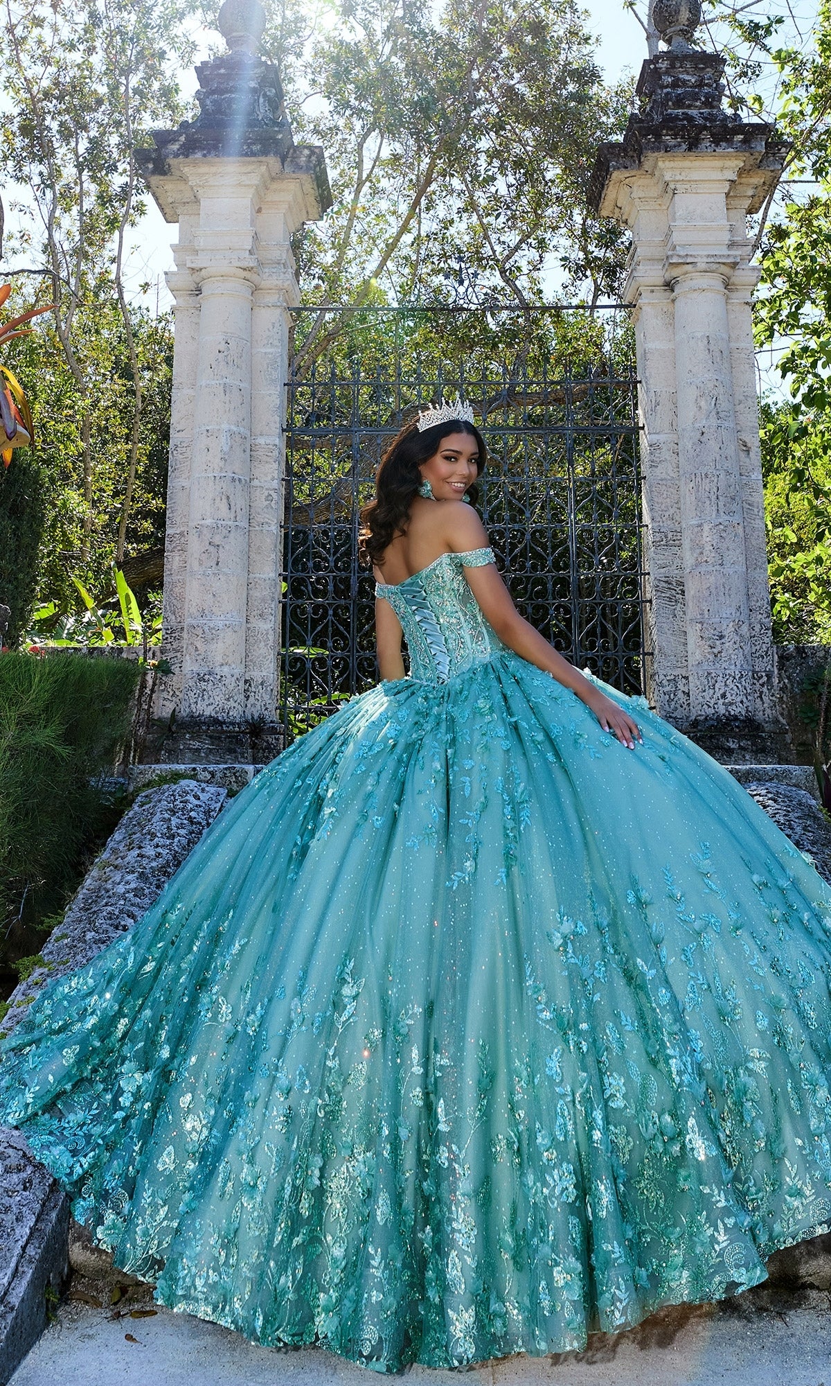 A young woman in the Princesa PR30131 Off-Shoulder Quinceanera Dress with 3D floral appliques and a corset bodice stands outdoors on stone steps amid greenery and pillars, facing slightly away.