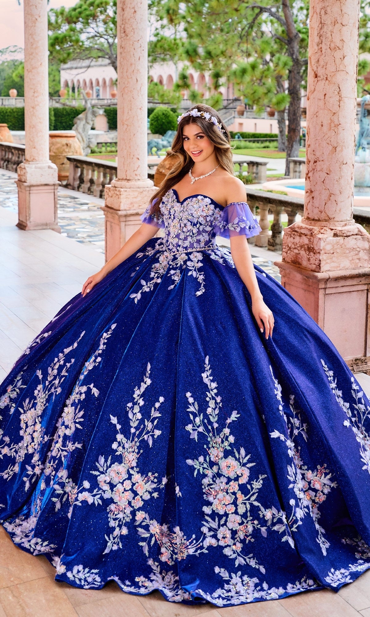 A woman stands in an outdoor corridor wearing the blue Quinceanera Dress PR30134 by Princesa, featuring a strapless corset bodice, floral embroidery, and a sparkling tiara.