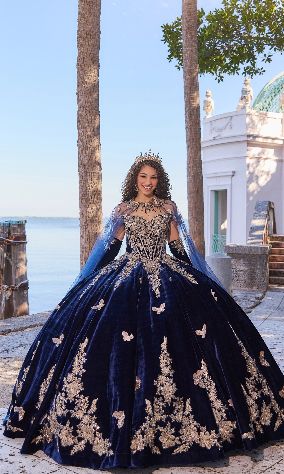 A woman stands outdoors by the water in the Princesa PR30136 velvet quinceanera dress with gold lace embroidery, a crown, and gloves, with trees and a white building in the background.