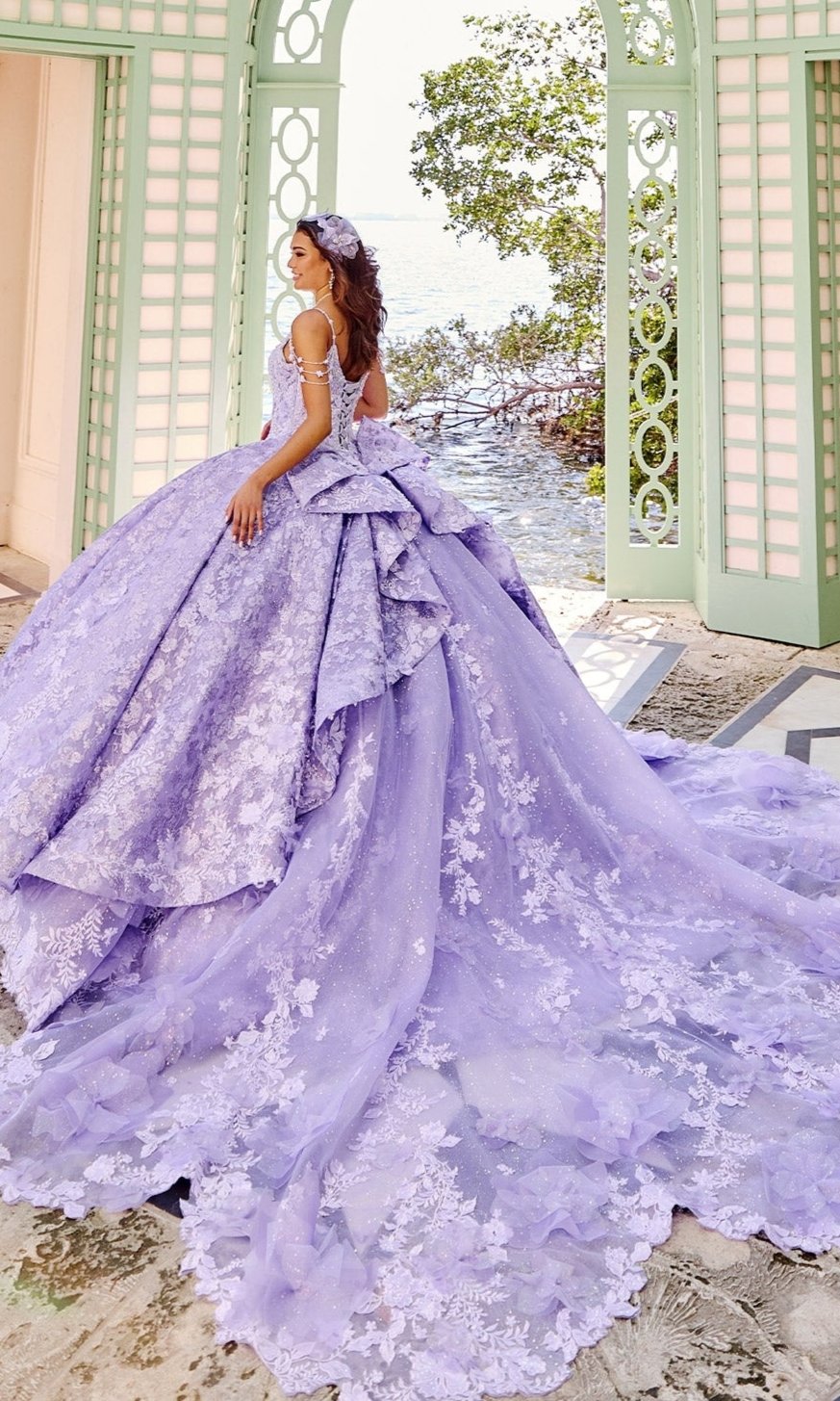 Wearing the Quinceanera Dress PR30139 by Princesa, a woman in a glitter tulle ball gown with lace details and a long train stands at open mint-green doors, looking back toward lush greenery outside.