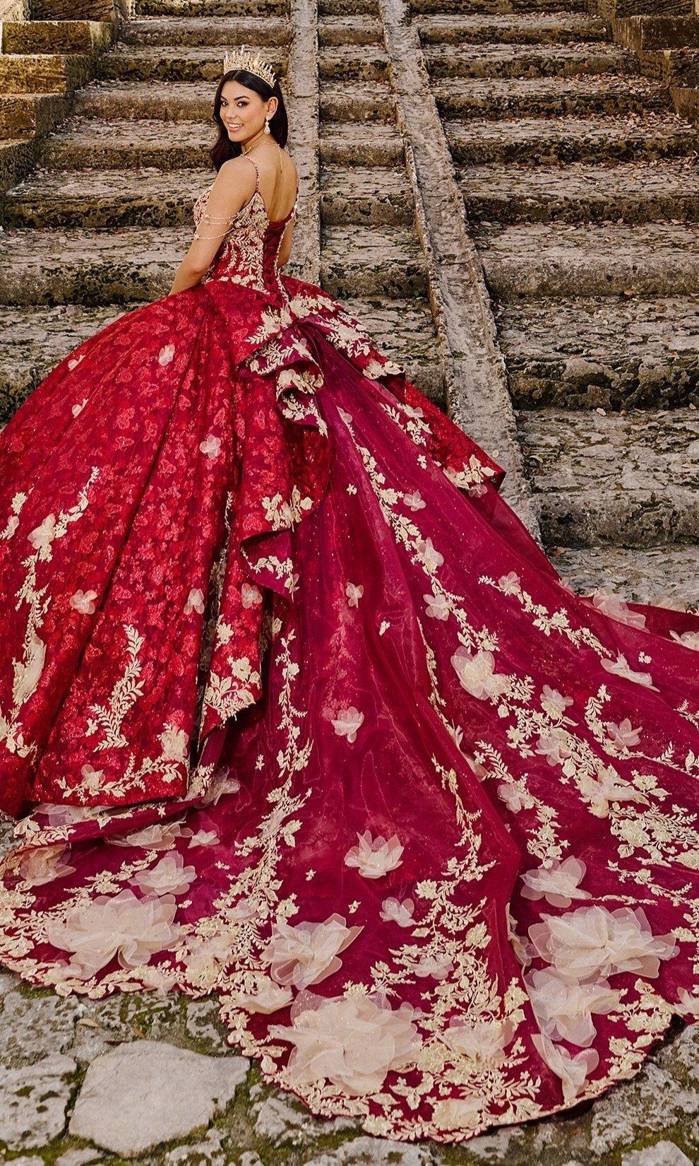 A woman in the Quinceanera Dress PR30139 By Princesa—an elaborate red gown with gold floral embroidery and a glitter tulle train—stands on stone steps, looking over her shoulder.