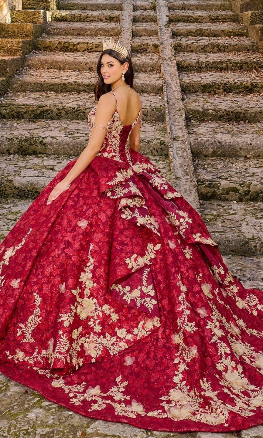 A woman stands on stone steps in the Quinceanera Dress PR30139 by Princesa, a red and gold ornate ball gown with glitter tulle, looking over her shoulder as her sparkling crown catches the light.