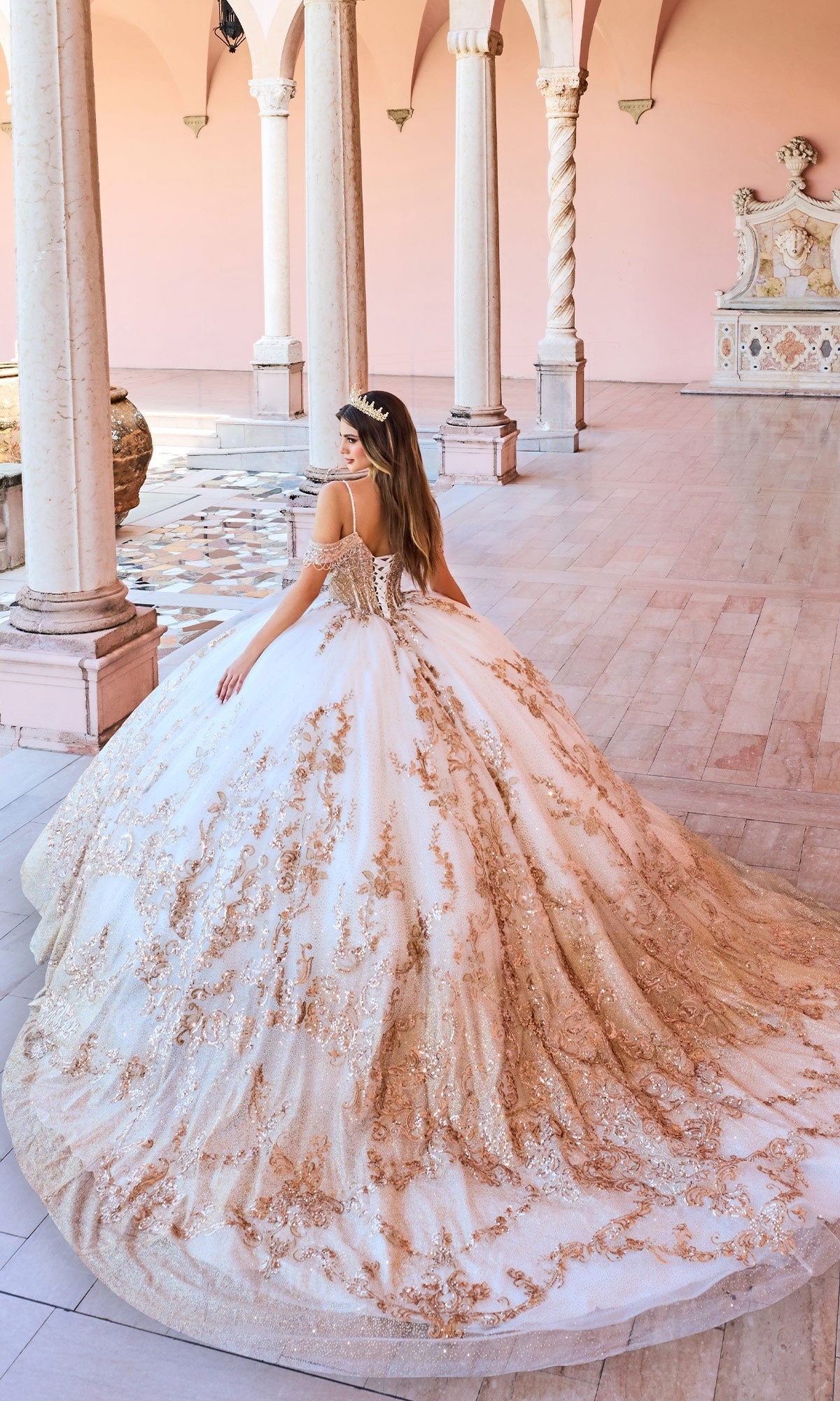 A woman stands in a marble hall wearing the Princesa PR30153 Lace Embroidered Quinceanera Dress, a grand white and gold gown with a corset bodice and long train, facing away to showcase its elegant detail.