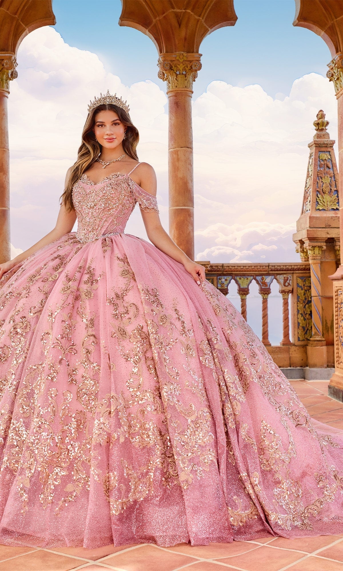 A young woman wears the Lace Embroidered Quinceanera Dress: Princesa PR30153, featuring a sparkling pink corset bodice, as she stands on a tiled terrace with ornate columns beneath a partly cloudy sky.