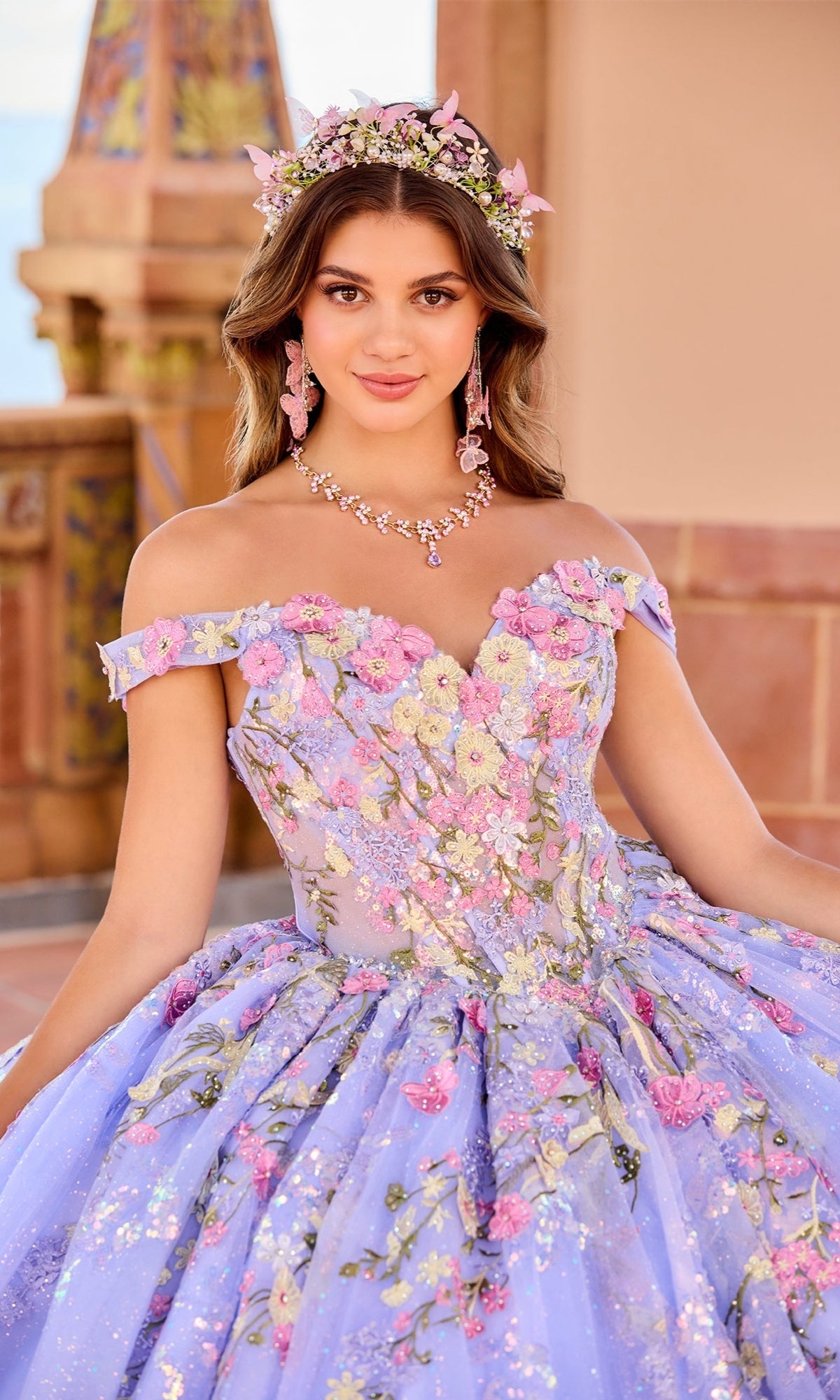 A young woman poses outdoors in the Quinceanera Dress PR30155 By Princesa, a lavender ball gown with 3D floral embroidery, off-the-shoulder sleeves, matching floral jewelry, and a flower crown.