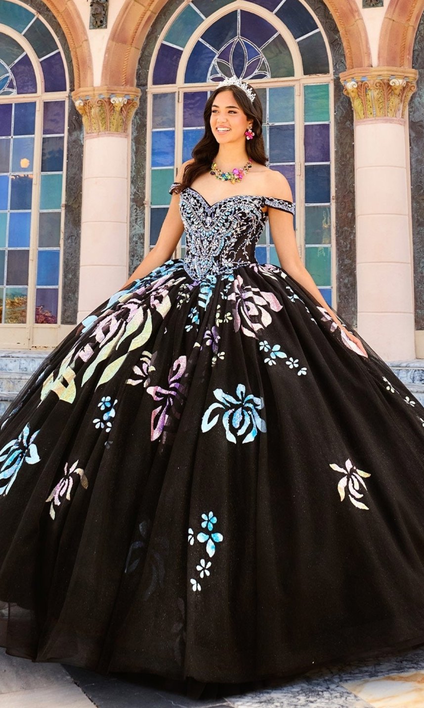 A woman in the PR30156 Quinceanera Dress by Princesa, featuring black fabric with colorful floral designs and detachable off-the-shoulder straps, stands before arched stained glass windows.