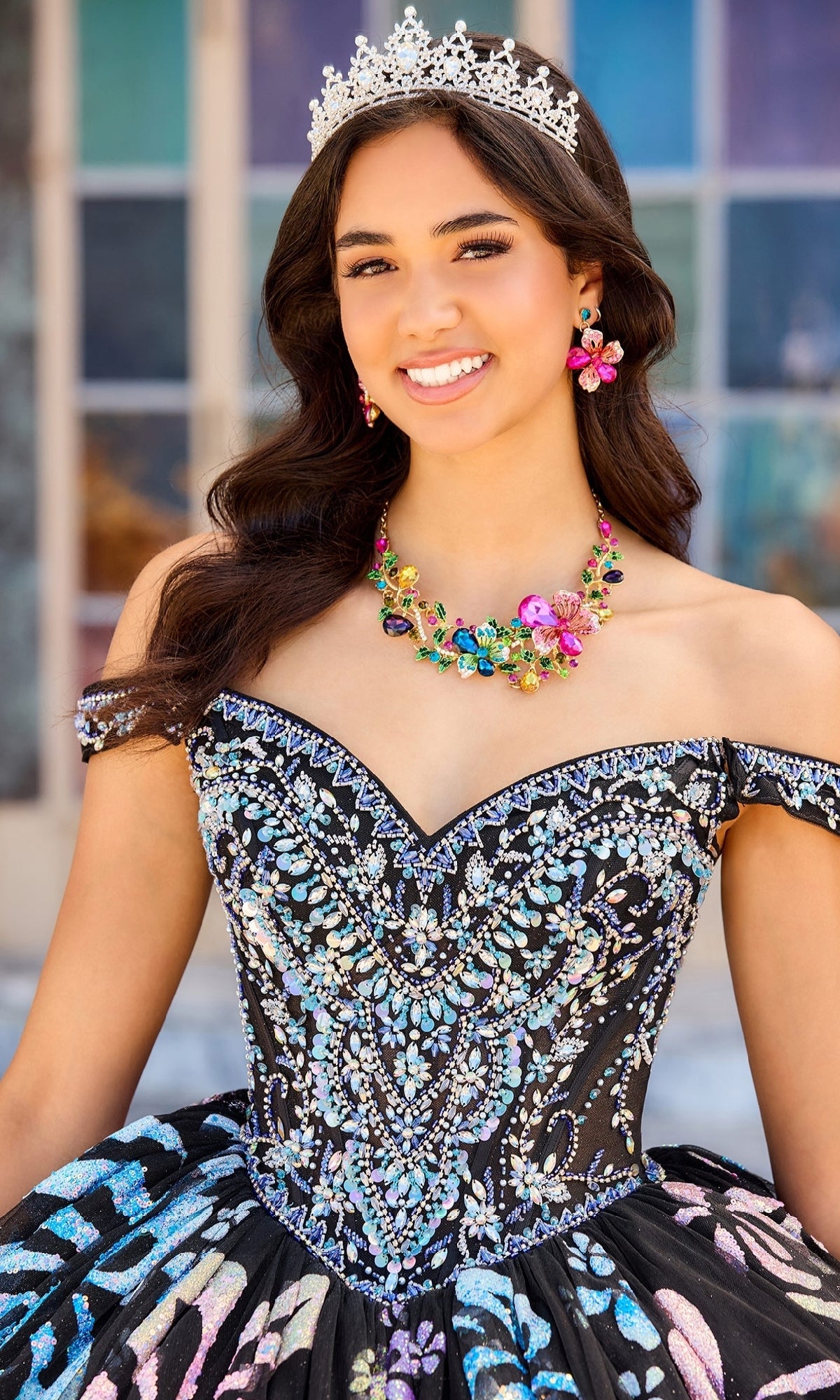 A young woman smiles outdoors wearing colorful earrings, a jeweled tiara, ornate floral necklace, and the Quinceanera Dress PR30156 By Princesa with beaded details and detachable off-the-shoulder straps.