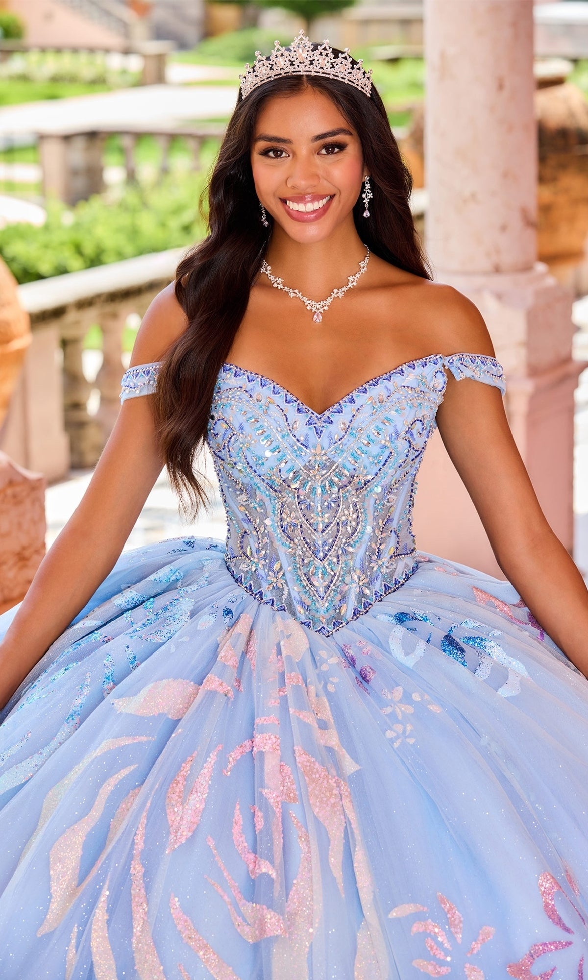 A young woman smiles outdoors near stone columns, wearing the Quinceanera Dress PR30156 By Princesa with sparkling blue fabric, detachable off-the-shoulder straps, a jeweled necklace, and tiara.