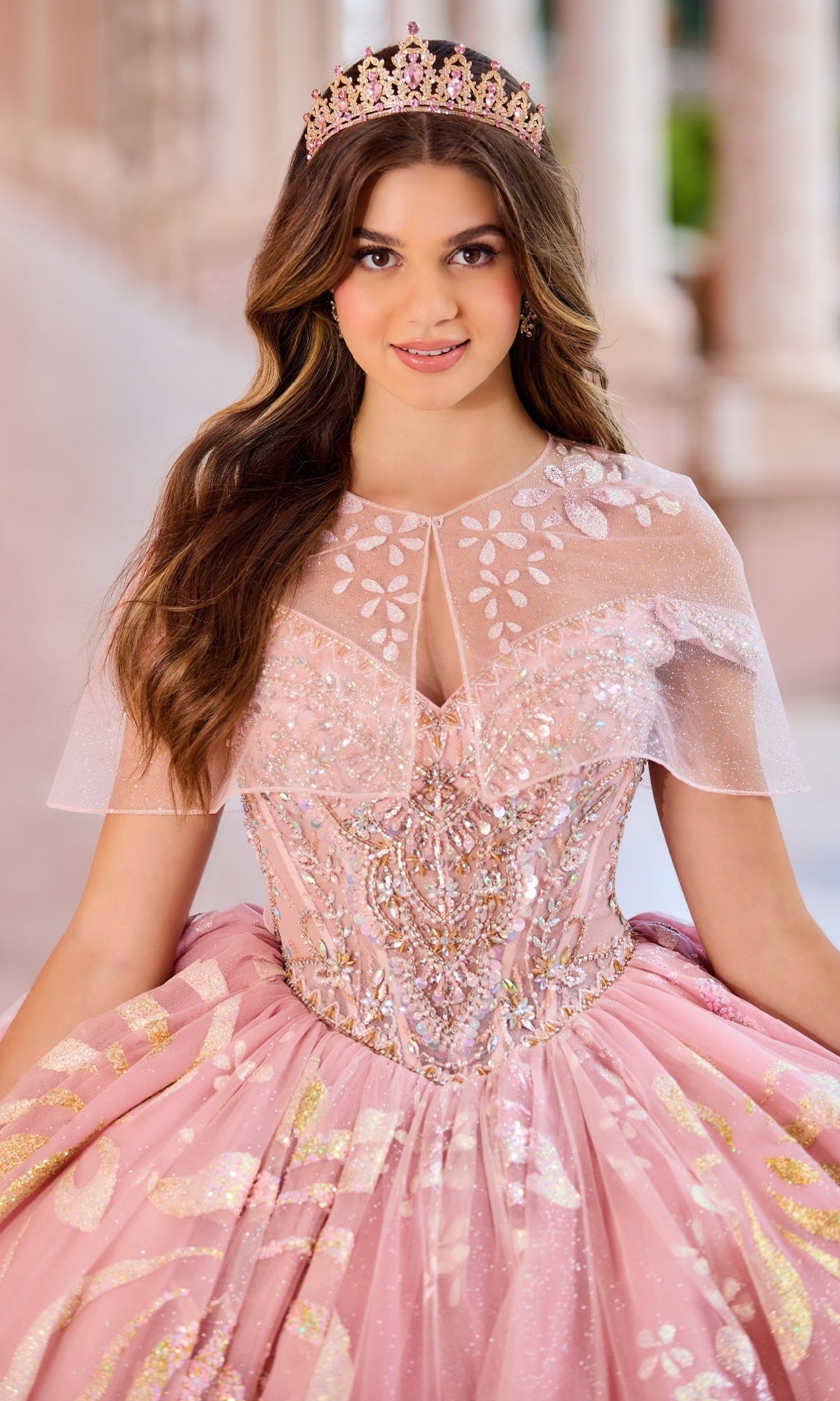 A young woman wears the Quinceanera Dress PR30156 By Princesa, featuring detachable off-the-shoulder straps and embellishments, paired with a tiara, posing outdoors before a blurred architectural backdrop.