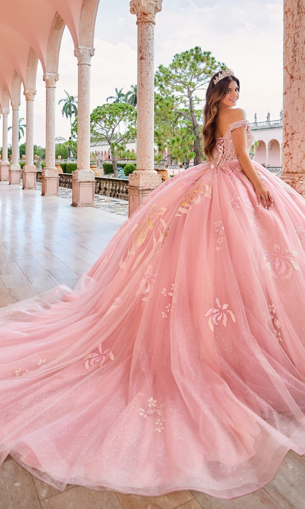 A woman in the Princesa Quinceanera Dress PR30156, featuring pink floral details and detachable off-the-shoulder straps, stands on a marble terrace with columns, trees, and a clear sky in the background.