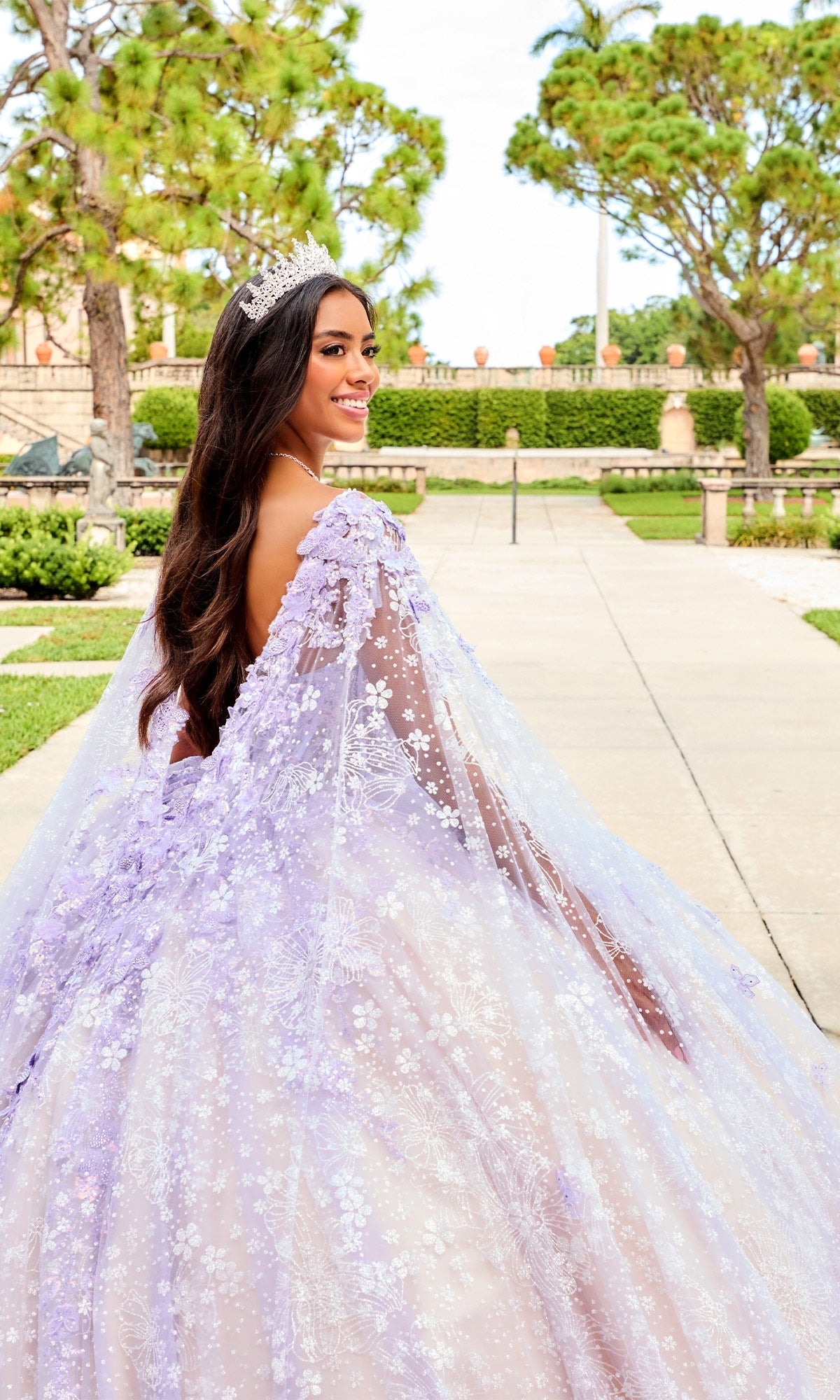 A woman in the Quinceanera Dress PR30158 By Princesa, a lavender floral gown with embroidered lace and sequins, stands outdoors on a walkway, smiling over her shoulder.