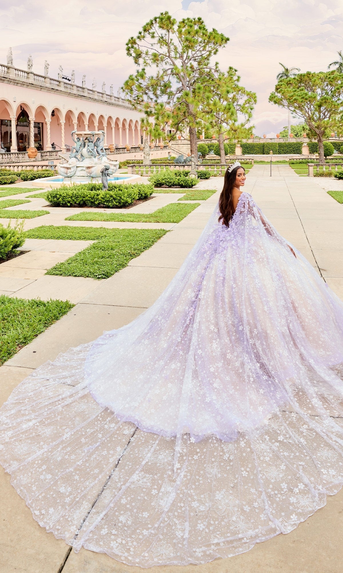 A woman wearing the Quinceanera Dress PR30158 by Princesa, a long lavender gown with embroidered lace, stands on a stone walkway in a formal garden with statues and manicured lawns.
