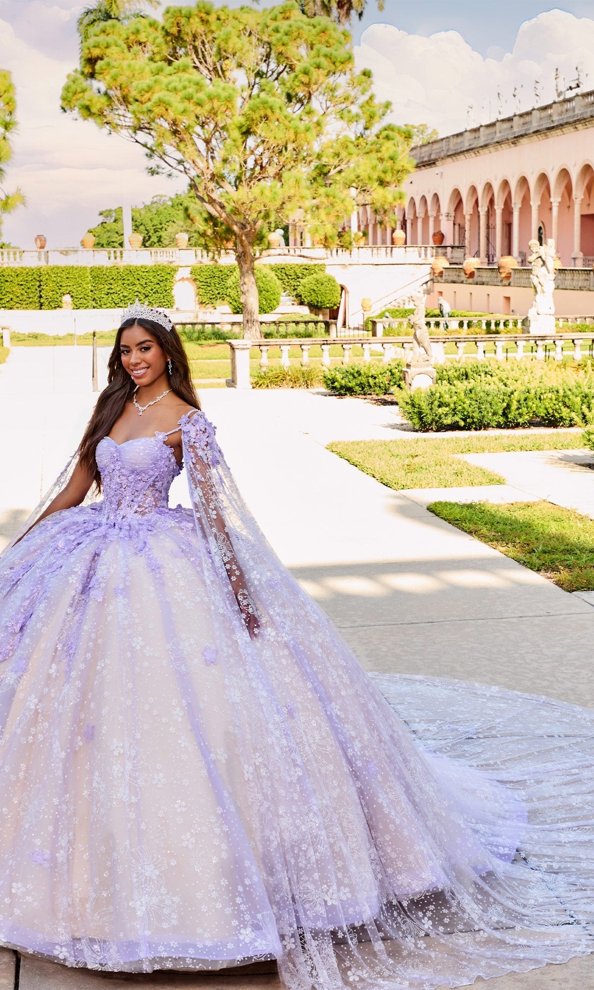 A young woman stands outdoors in a formal garden near an arched, statue-adorned building, wearing the shimmering Quinceanera Dress PR30158 By Princesa—a lavender gown with a lace cape and delicate embroidered details.
