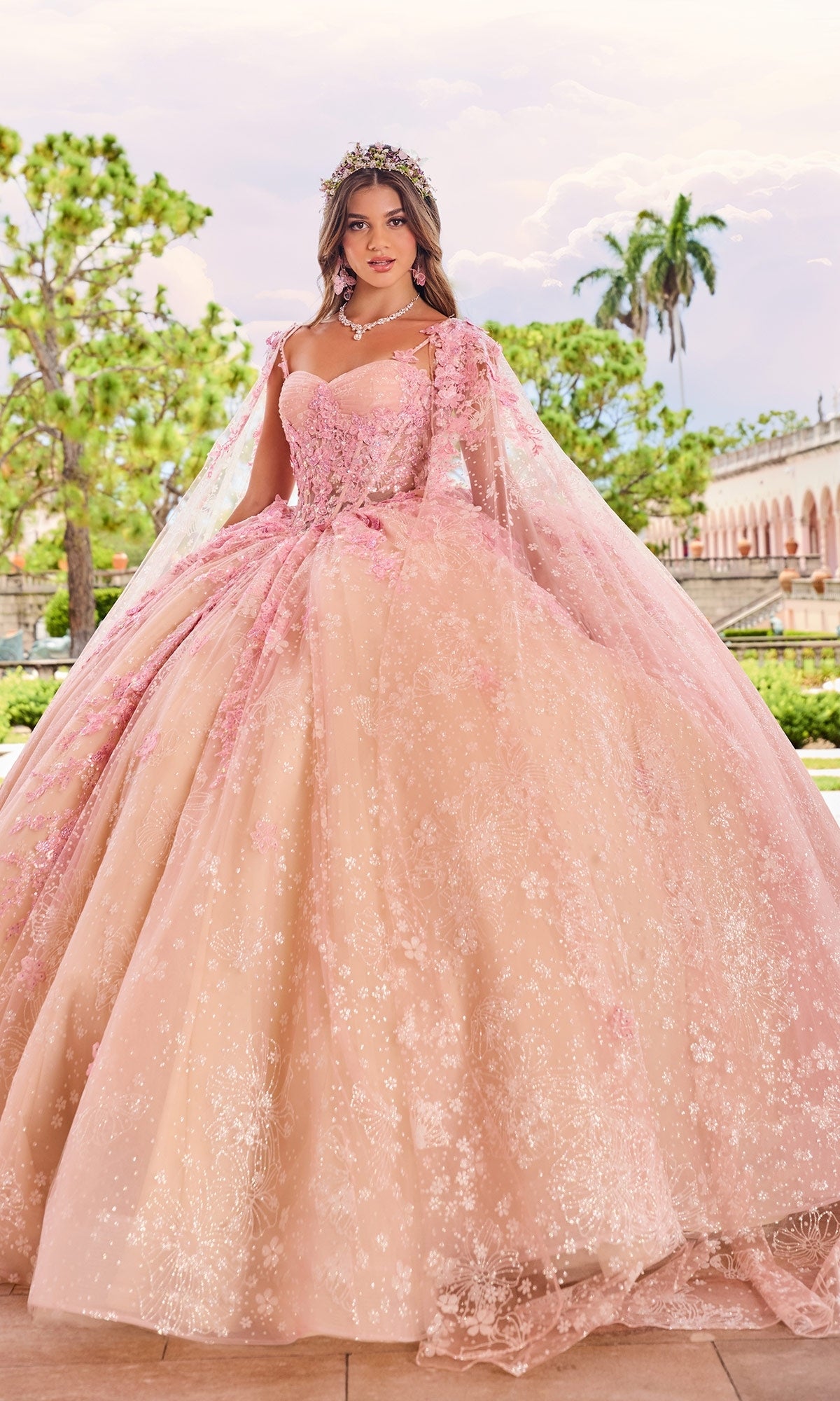 A woman wearing the Quinceanera Dress PR30158 By Princesa, featuring pink floral embroidery, lace details, a matching cape, and tiara stands near a garden with classical architecture.