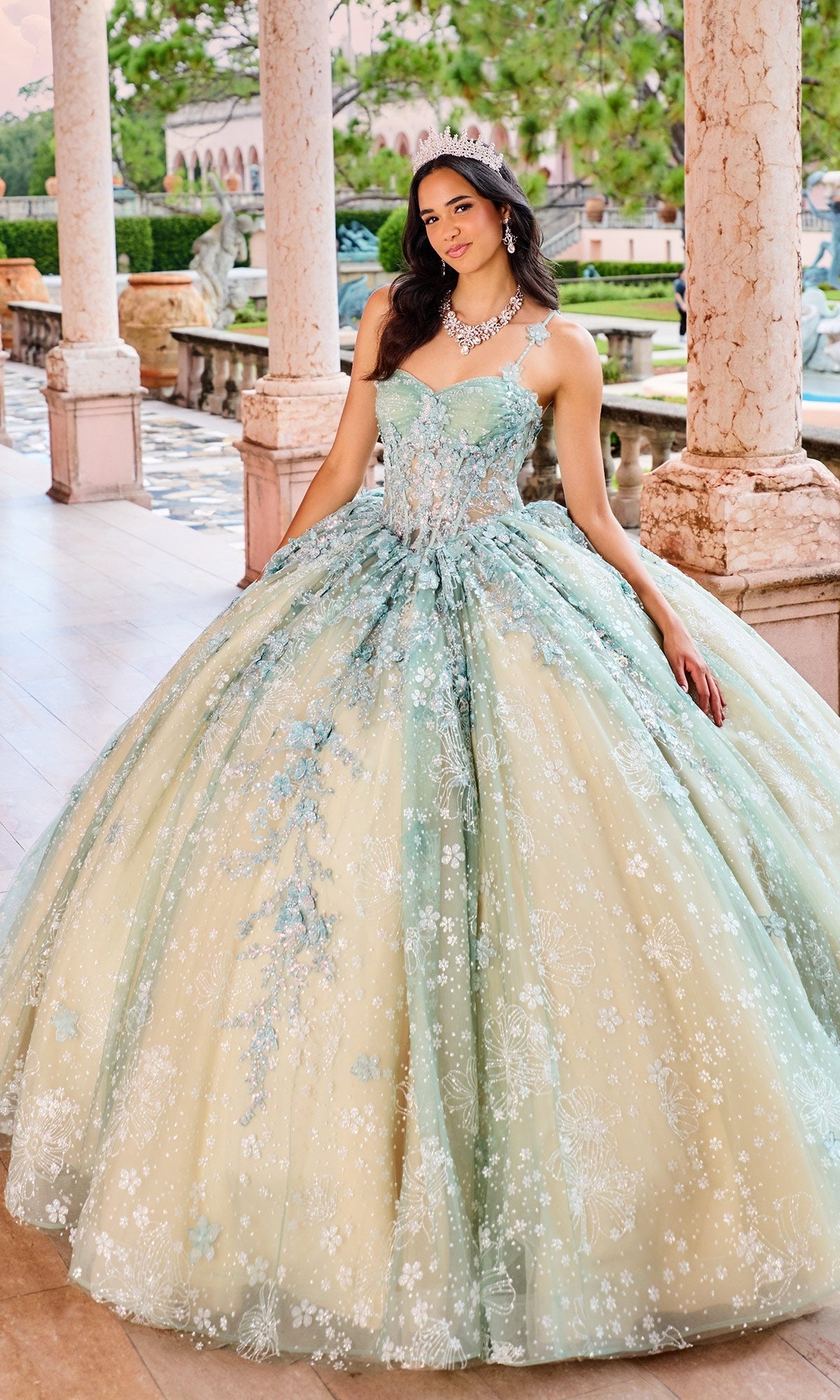 A woman in the Quinceanera Dress PR30158 By Princesa, featuring ornate pastel blue and gold lace with floral details, stands outdoors by stone columns, accessorized with a tiara and necklace.