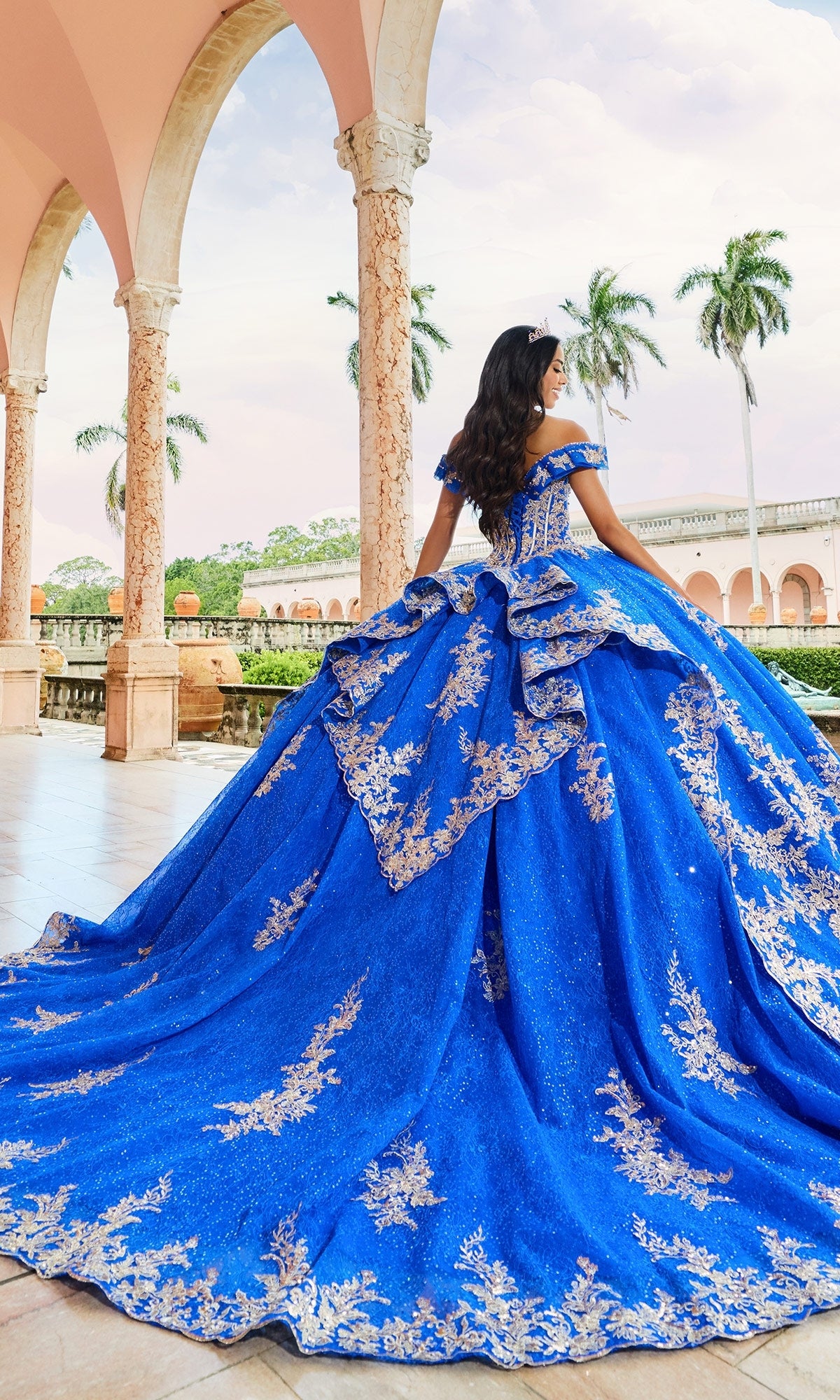 A woman wearing the Quinceanera Dress PR30163 by Princesa, a lace ball gown with blue and gold embellishments, stands outdoors under arches with palm trees in the background, facing away.