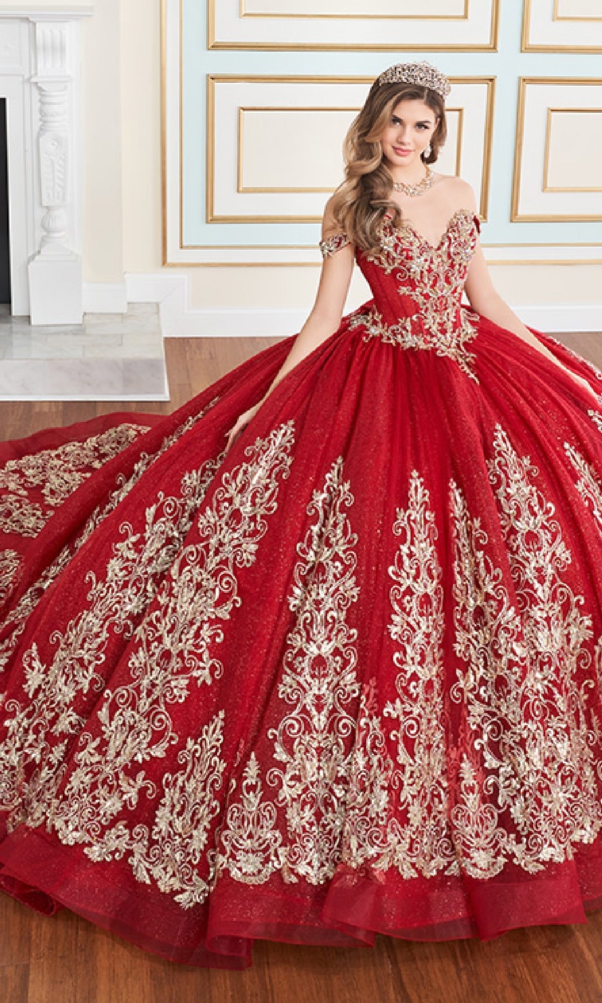 A woman wears the Quinceanera Dress PR30171 By Princesa, a red ball gown with gold embroidery and layers of glitter tulle, paired with a tiara. She sits indoors on a wooden floor before a decorated wall and fireplace.