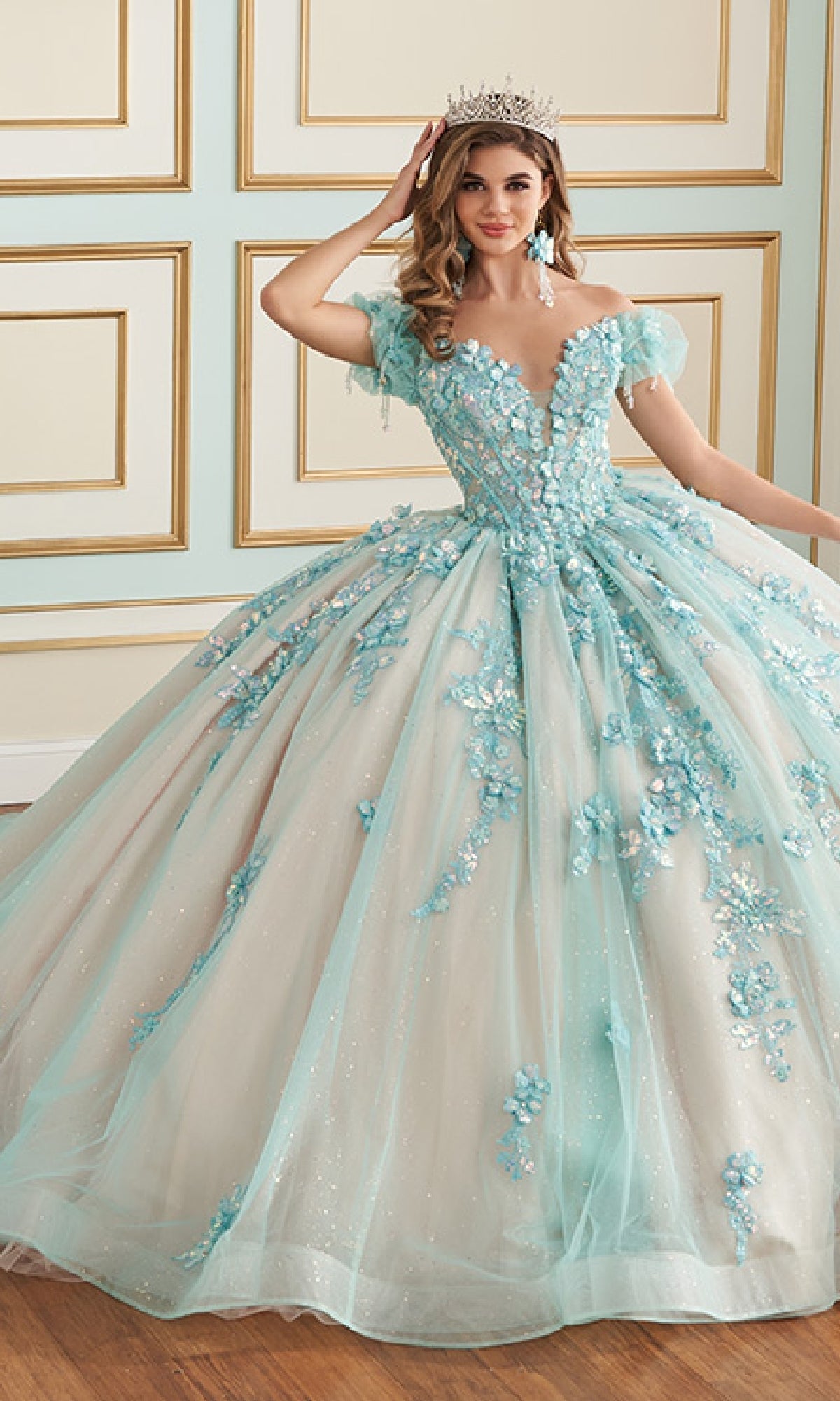 A young woman wears the Quinceanera Dress PR30174 By Princesa, featuring 3D floral appliques and a crown, as she poses in an elegant room with paneled walls.
