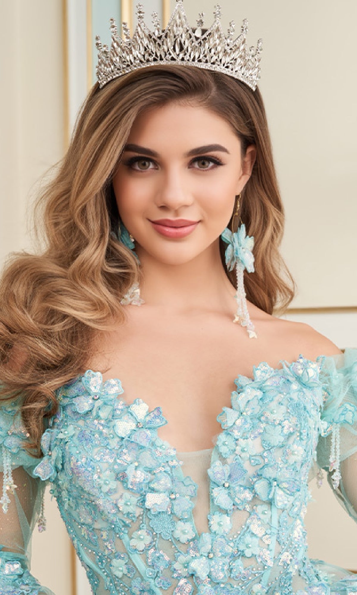 A woman wearing the Quinceanera Dress PR30174 By Princesa, a light blue ball gown with off-the-shoulder sleeves and 3D floral appliques, smiles at the camera.