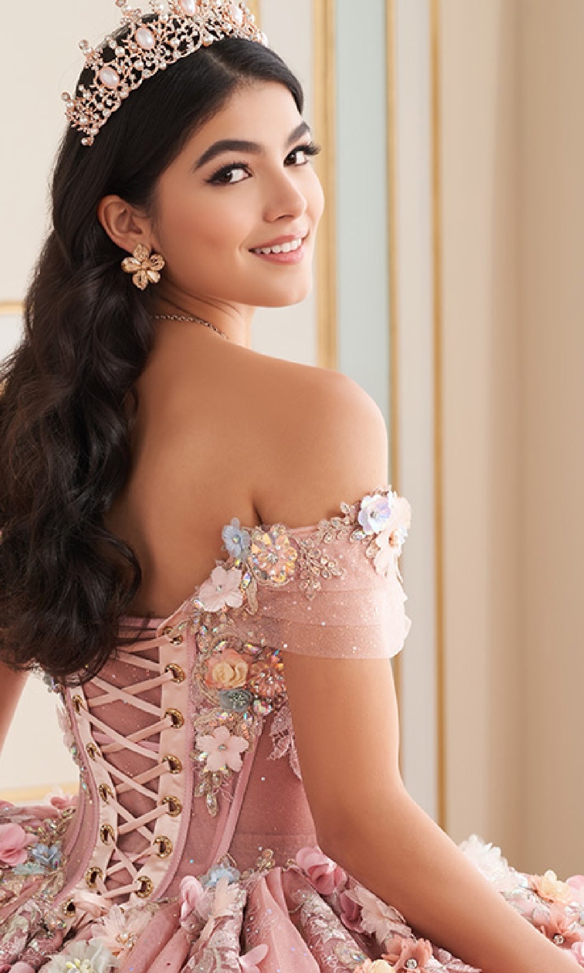 A young woman wearing the Quinceanera Dress PR30175 by Princesa—pink with 3D florals, stone accents, and a tiara—smiles over her shoulder while seated in a formal setting.