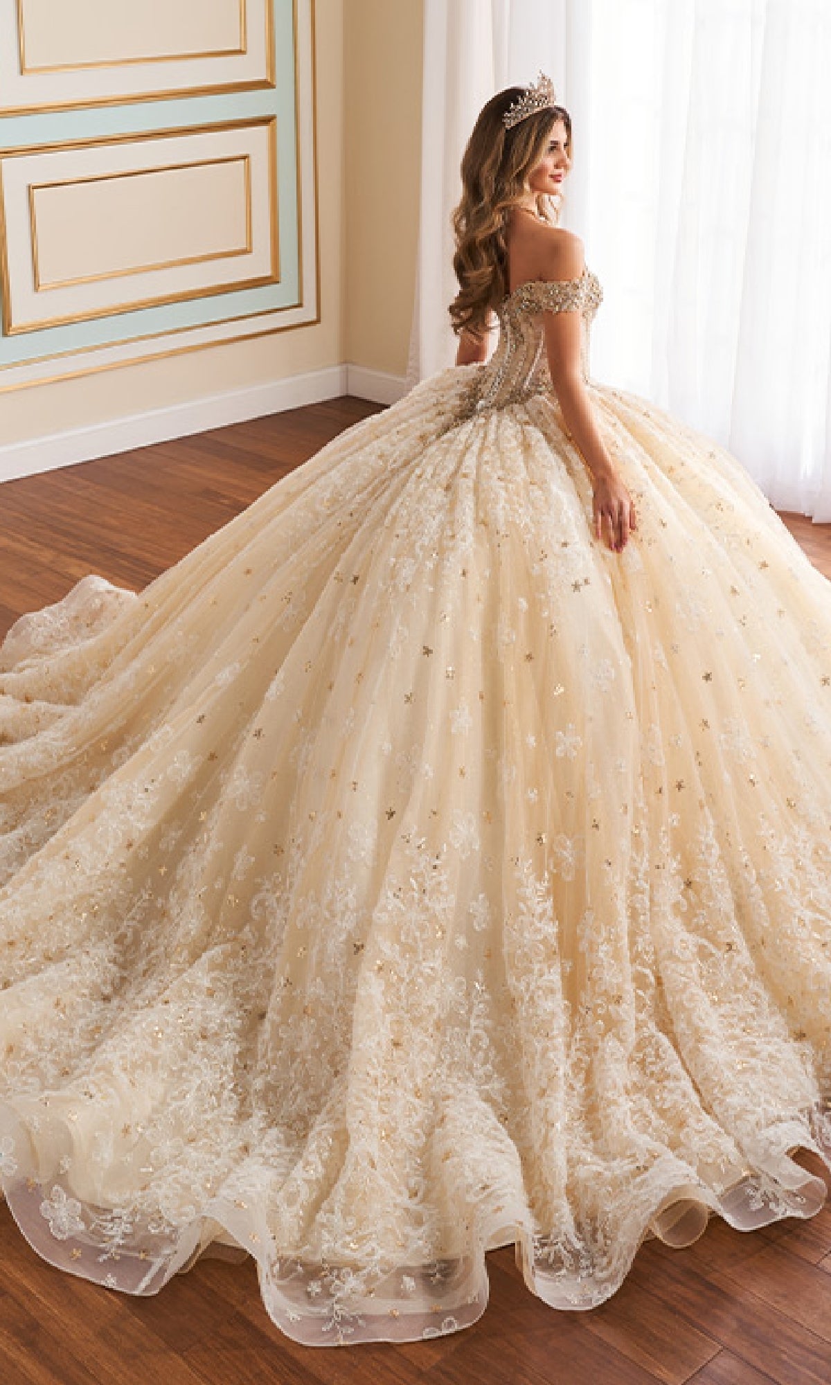 A woman smiles and stands on a wooden floor wearing the Quinceanera Dress PR30177 By Princesa, an ornate off-the-shoulder gown with a corset bodice and lace embroidery. Her tiara catches the light as she faces slightly away.