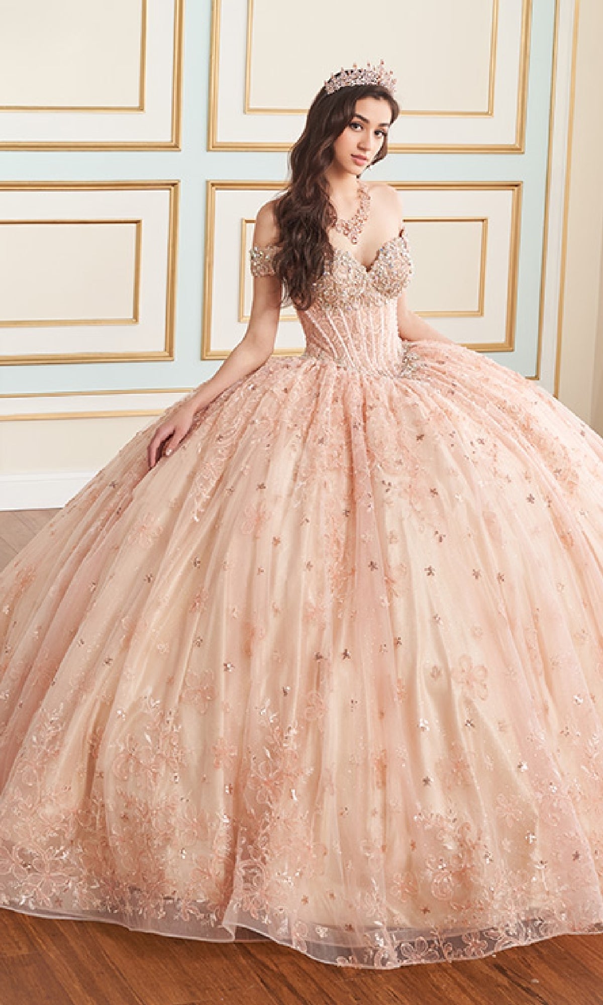 A woman sits indoors against a decorated wall wearing the Quinceanera Dress PR30177 By Princesa, featuring an off-the-shoulder corset bodice with pink embellishments, and finishes her look with a sparkling tiara.