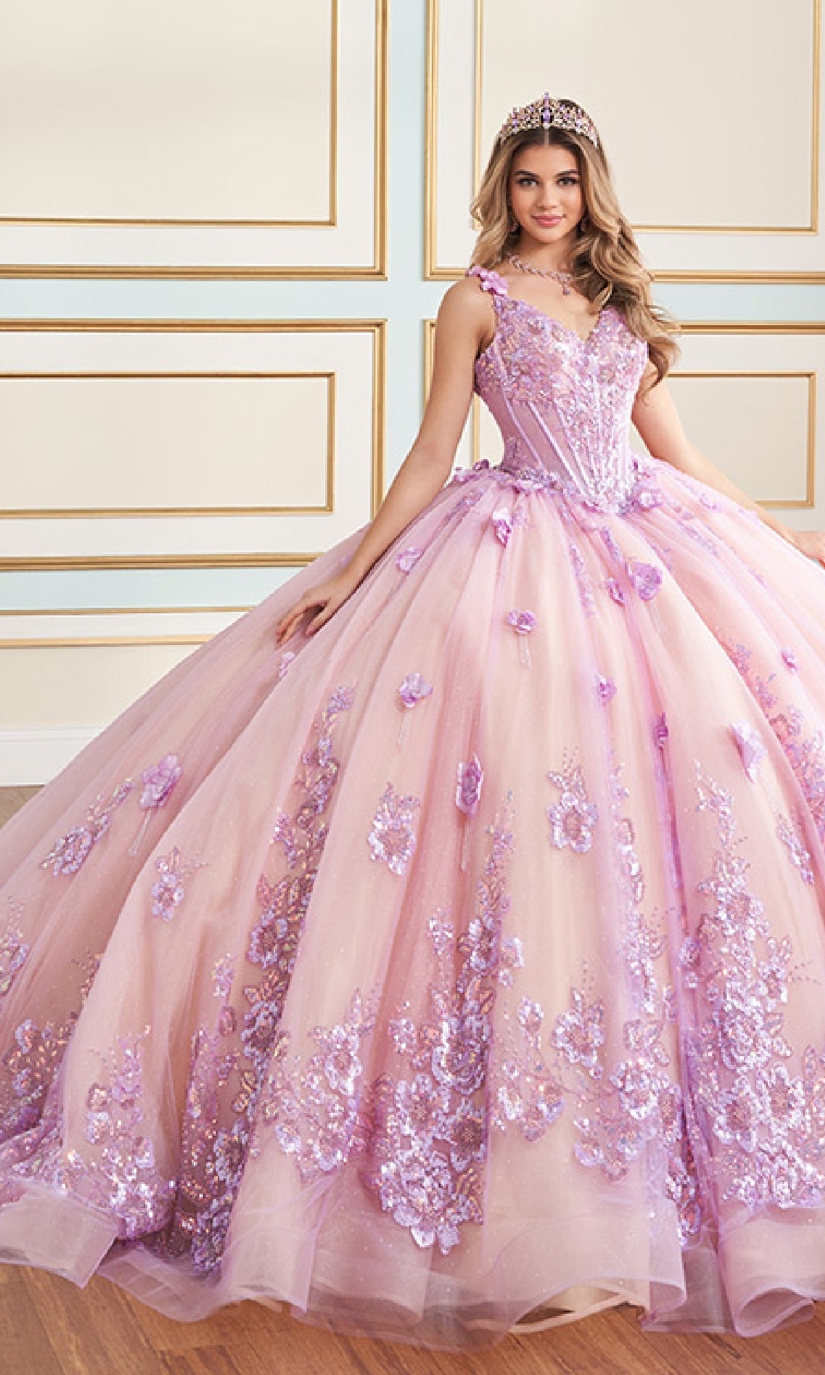 A young woman in the Quinceanera Dress PR30179 By Princesa, a pink and lavender glitter tulle gown with 3D flowers and a tiara, stands indoors against a white and gold wall.