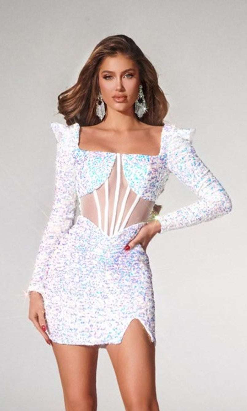 A woman poses in the Portia and Scarlett PS22555 short prom dress, featuring sequins, sheer paneling, long sleeves, silver heels, and statement earrings against a plain background.