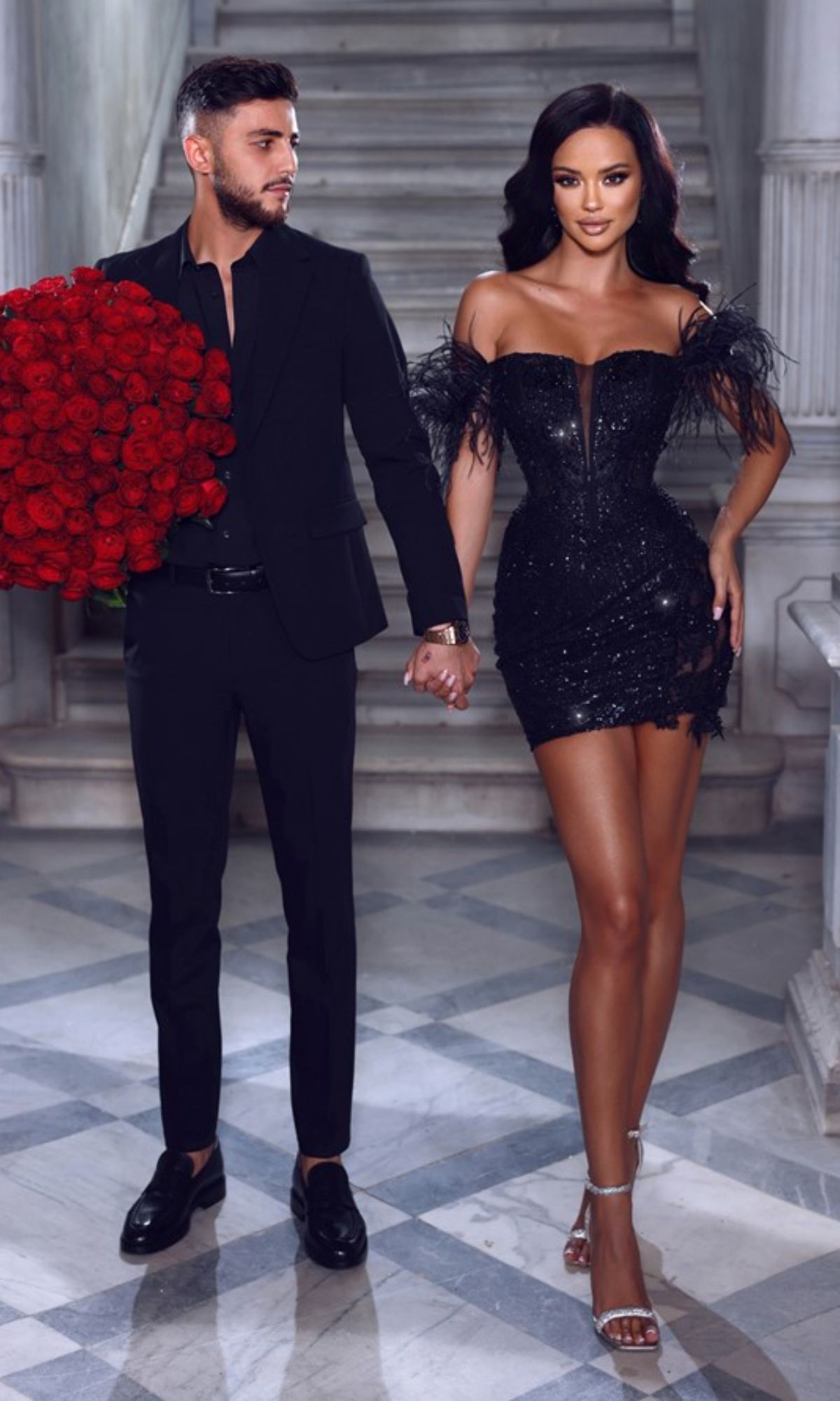 A man in a black suit holds a large bouquet of red roses and the hand of a woman wearing the Short Party Dress PS23147 by Portia and Scarlett in black, featuring feather details, as they stand on marble steps.