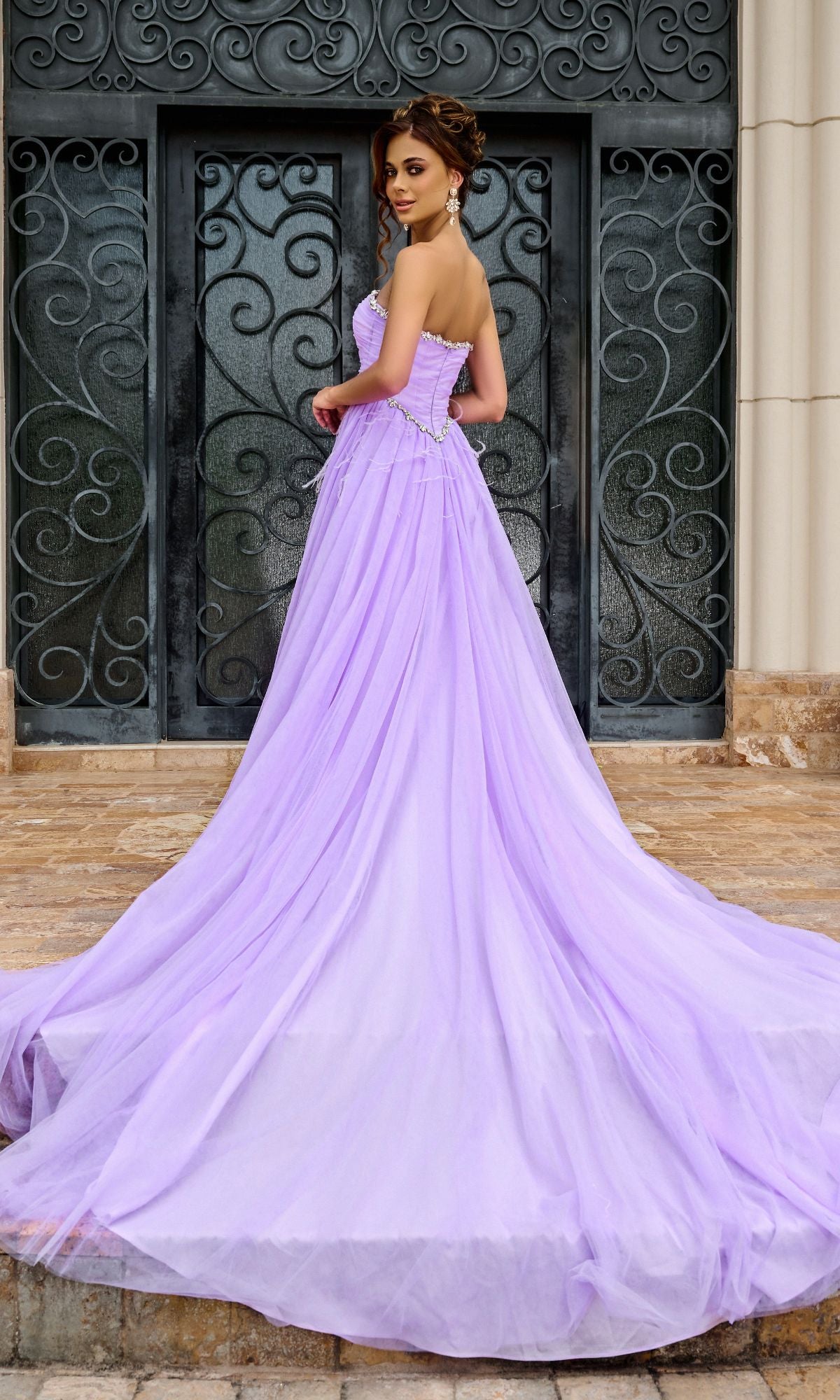 A woman wearing the Long Prom Dress PS24632 by Portia and Scarlett, featuring a strapless tulle A-line silhouette with a beaded sweetheart neckline and long train, stands on a stone path before an ornate black door.