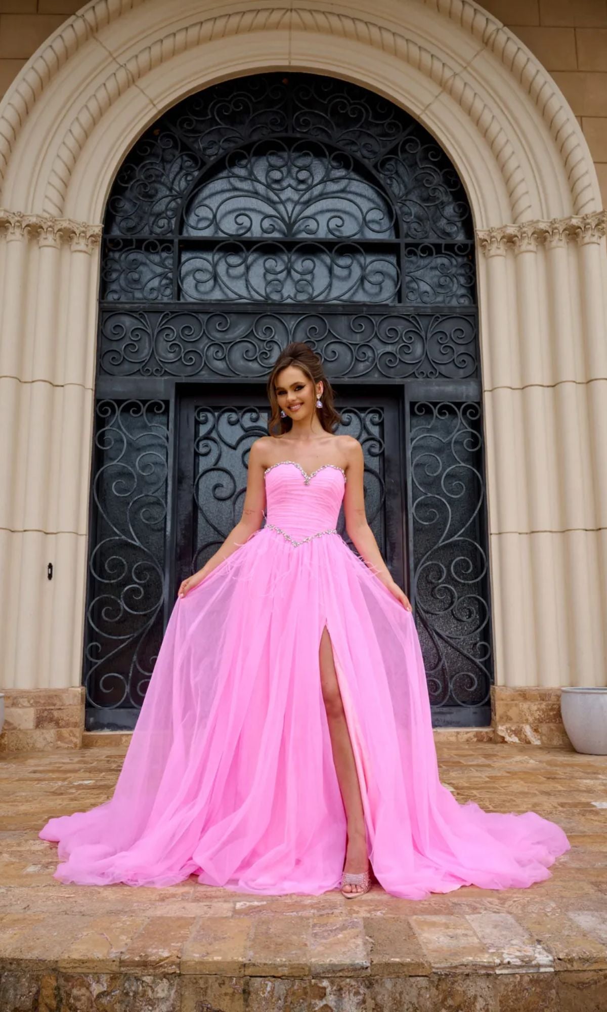 A woman stands before a grand black door, wearing the Long Prom Dress PS24632 by Portia and Scarlett, a strapless A-line tulle ball gown with a beaded sweetheart neckline, floor-length pink skirt, and thigh-high slit.