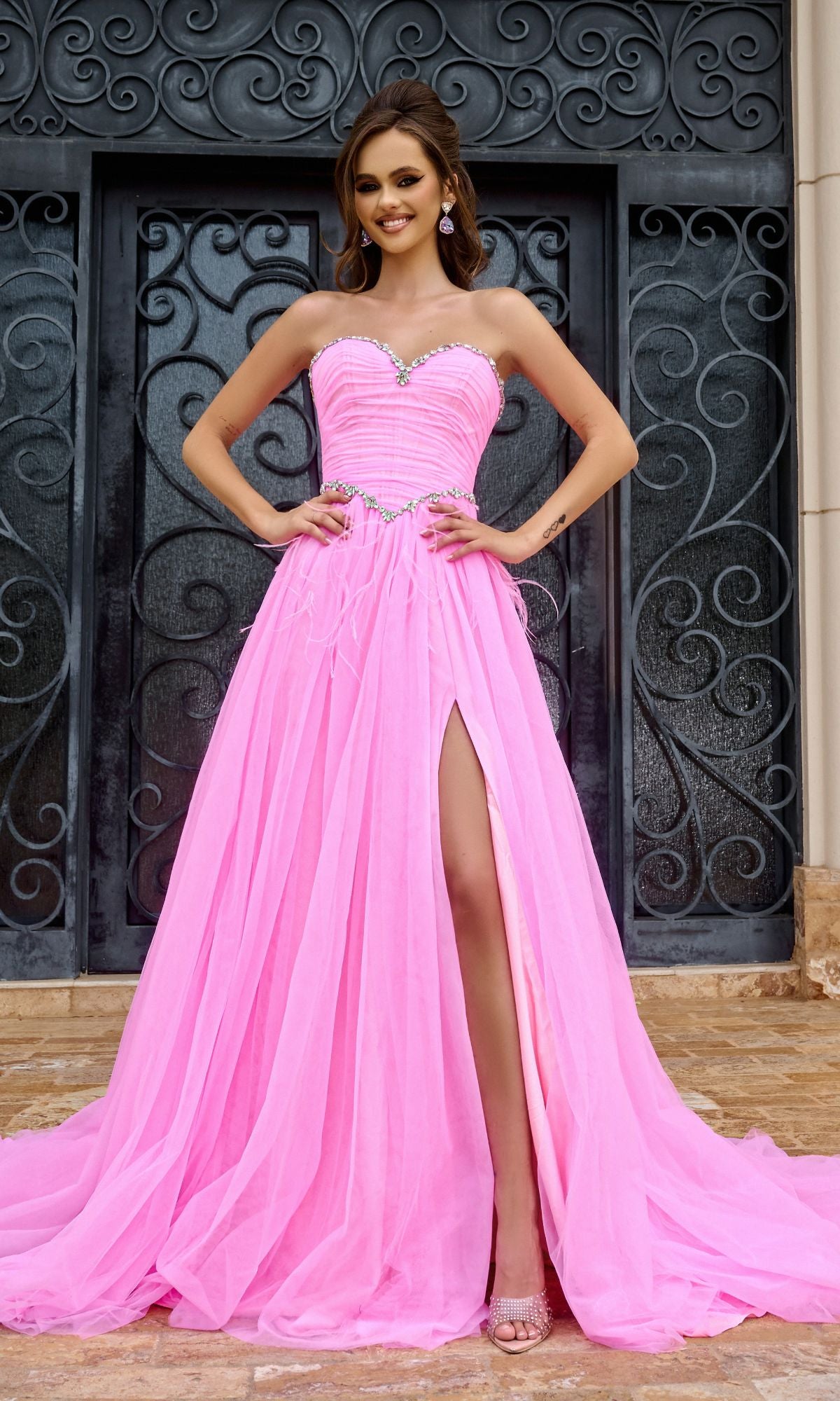 A woman smiles in front of ornate black doors, wearing the Long Prom Dress PS24632 by Portia and Scarlett—a pink, strapless, floor-length gown with a beaded sweetheart neckline and high slit.