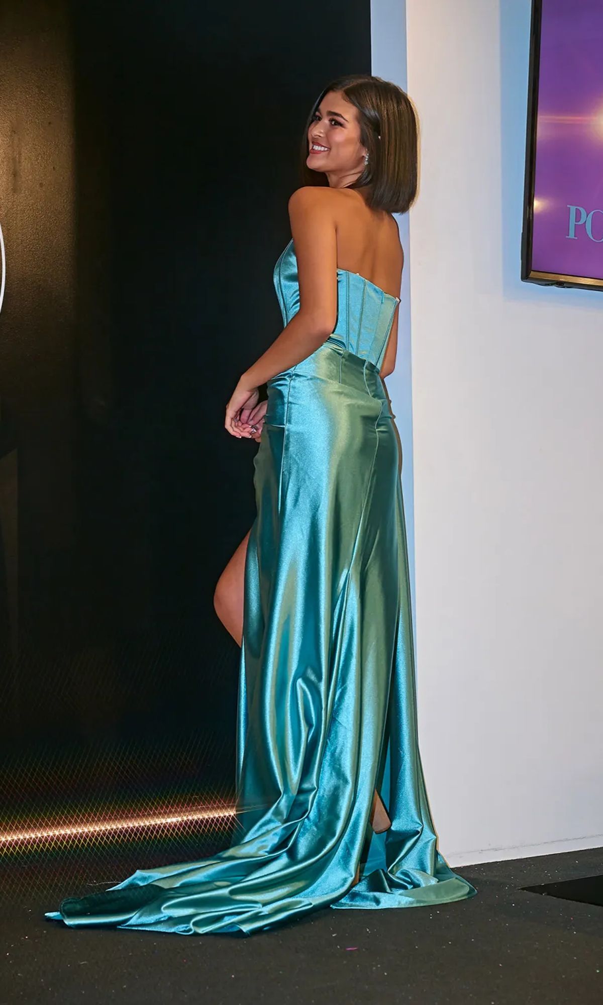 A woman wearing the Portia & Scarlett PS24682 Strapless Satin Prom Gown with a sweeping train stands indoors, smiling over her shoulder.