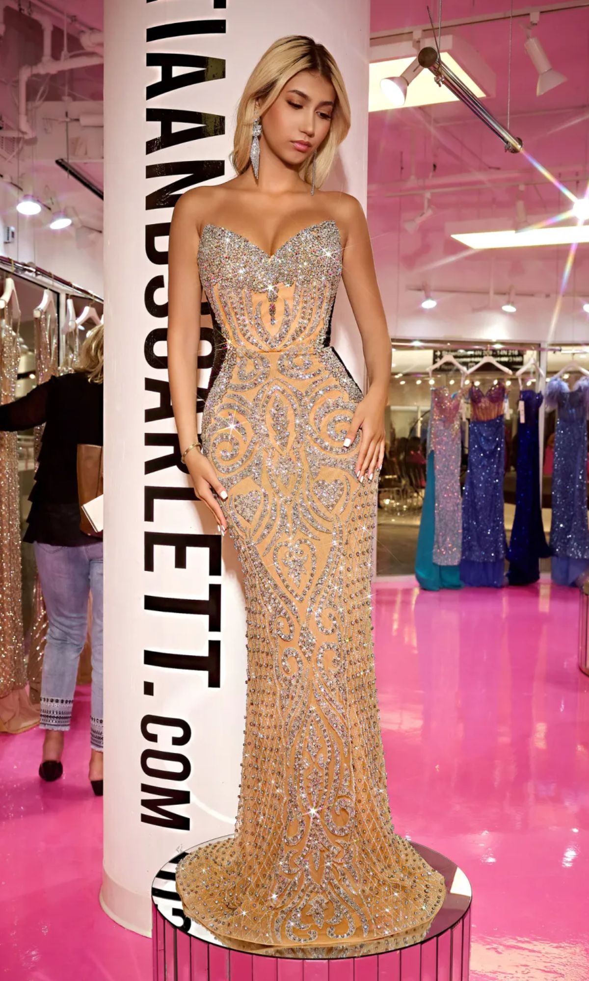 A woman wears the Long Prom Dress PS24867C by Portia and Scarlett in Nude/Silver/Black, standing in a brightly lit boutique with pink floors and evening gowns displayed in the background.