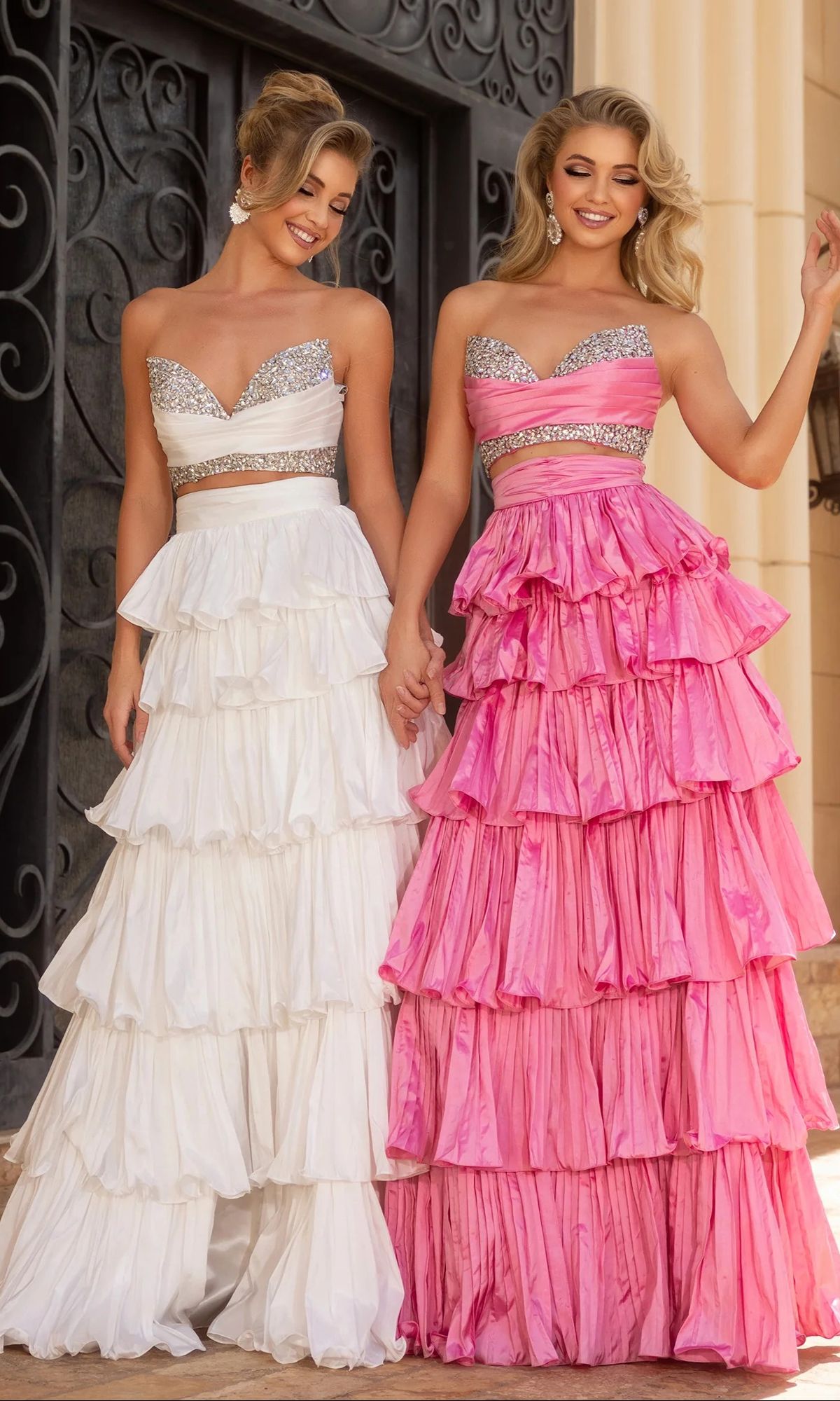Two women hold hands, each wearing a Portia and Scarlett PS25164 long prom dress—one in white and silver, the other in pink and silver—both featuring sparkly embellishments as they pose before an ornate black door.