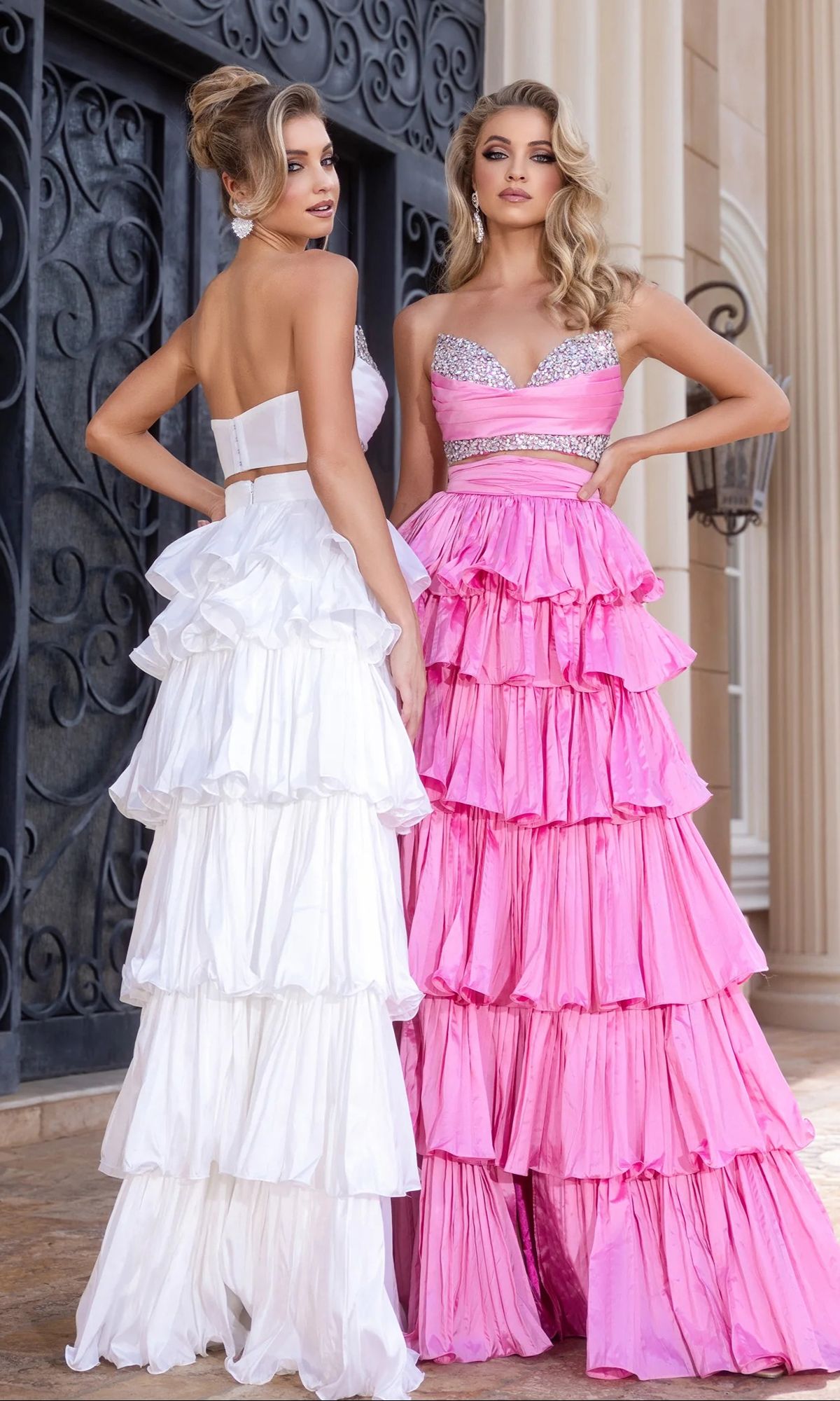 Two women pose outdoors before ornate doors and columns, each wearing the Portia and Scarlett PS25164 long prom dress—one in white, one in pink. The dresses feature tiered designs with subtle sparkly embellishments for an elegant look.