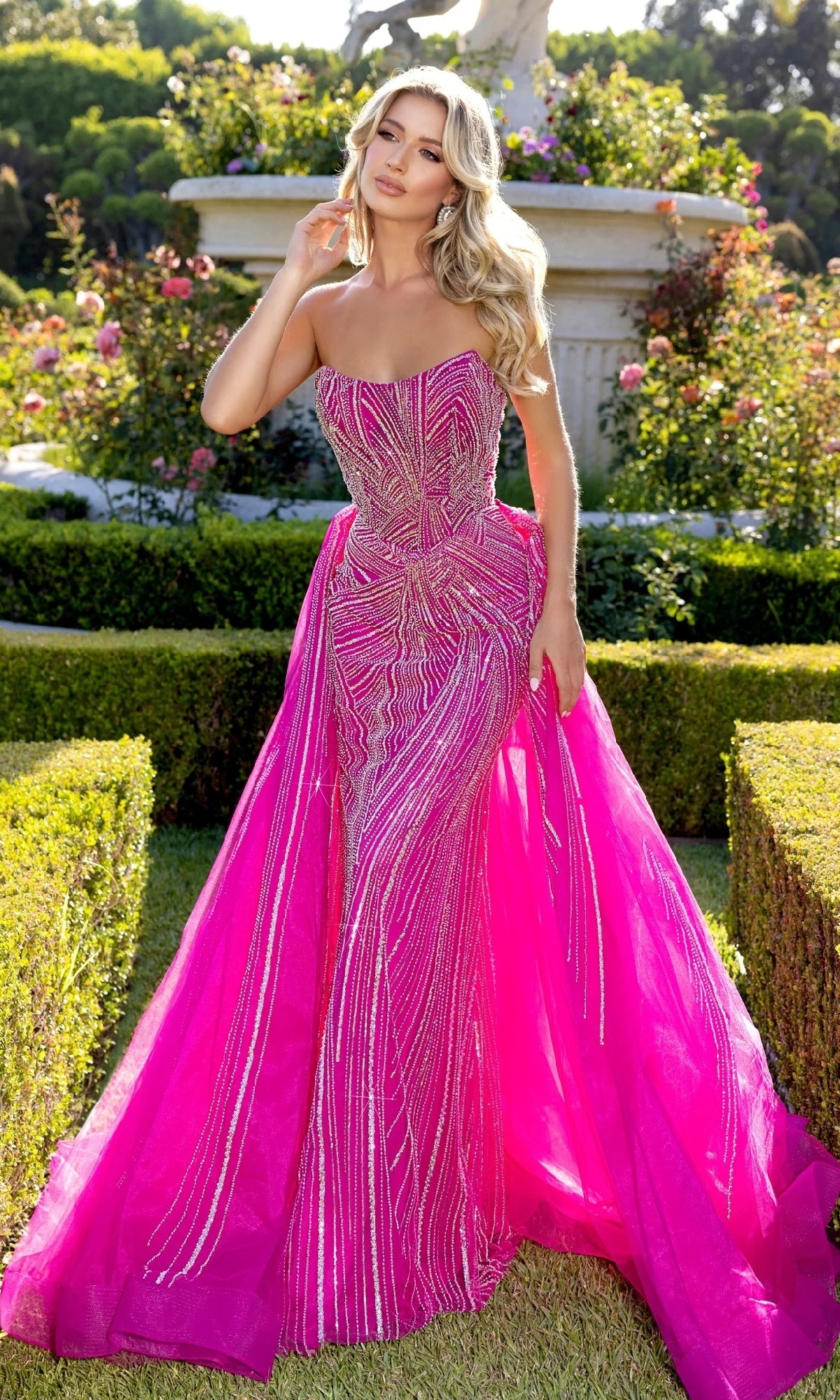 A woman stands outdoors in a manicured garden wearing the Portia and Scarlett PS25175 Long Prom Dress—a dramatic fuchsia gown with silver sequin details and a voluminous strapless tulle skirt.