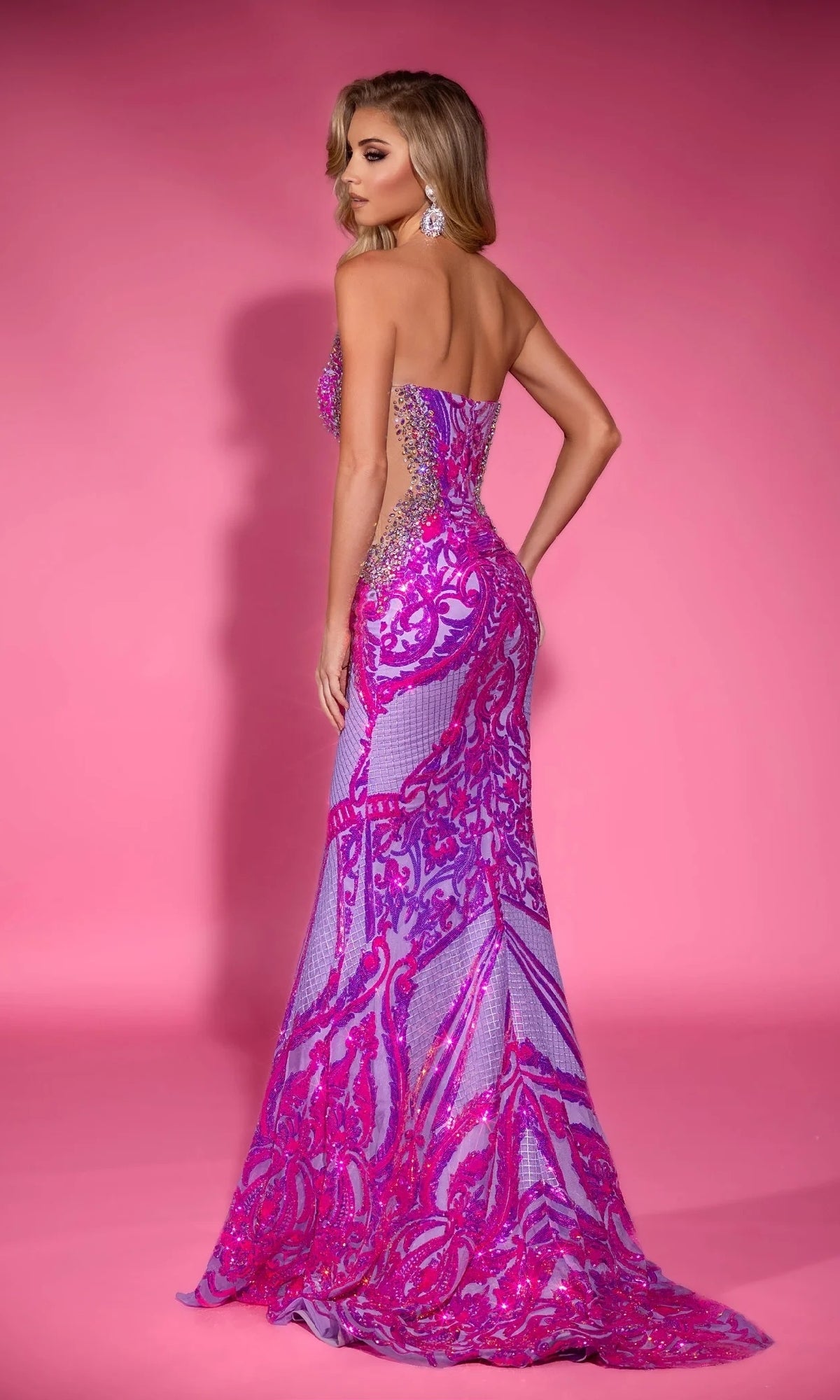 A woman wears the Portia and Scarlett PS25263 long prom dress, featuring purple and pink patterned sequins, as she stands against a pink background, glancing over her shoulder.