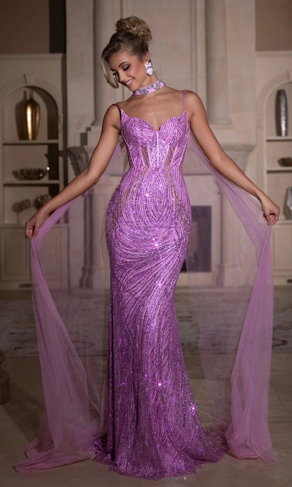 A woman smiles indoors, posing in the Portia and Scarlett PS25266 long prom dress—a sparkling lavender sequin gown with sheer panels and a tulle cape that she holds out to the sides.