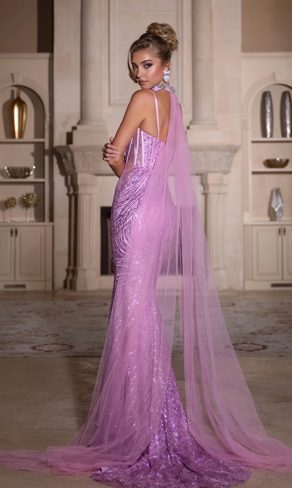 A woman stands indoors in a Long Prom Dress: Portia and Scarlett PS25266, a floor-length sequin gown with a sheer tulle cape detail, looking over her shoulder in an elegant setting.