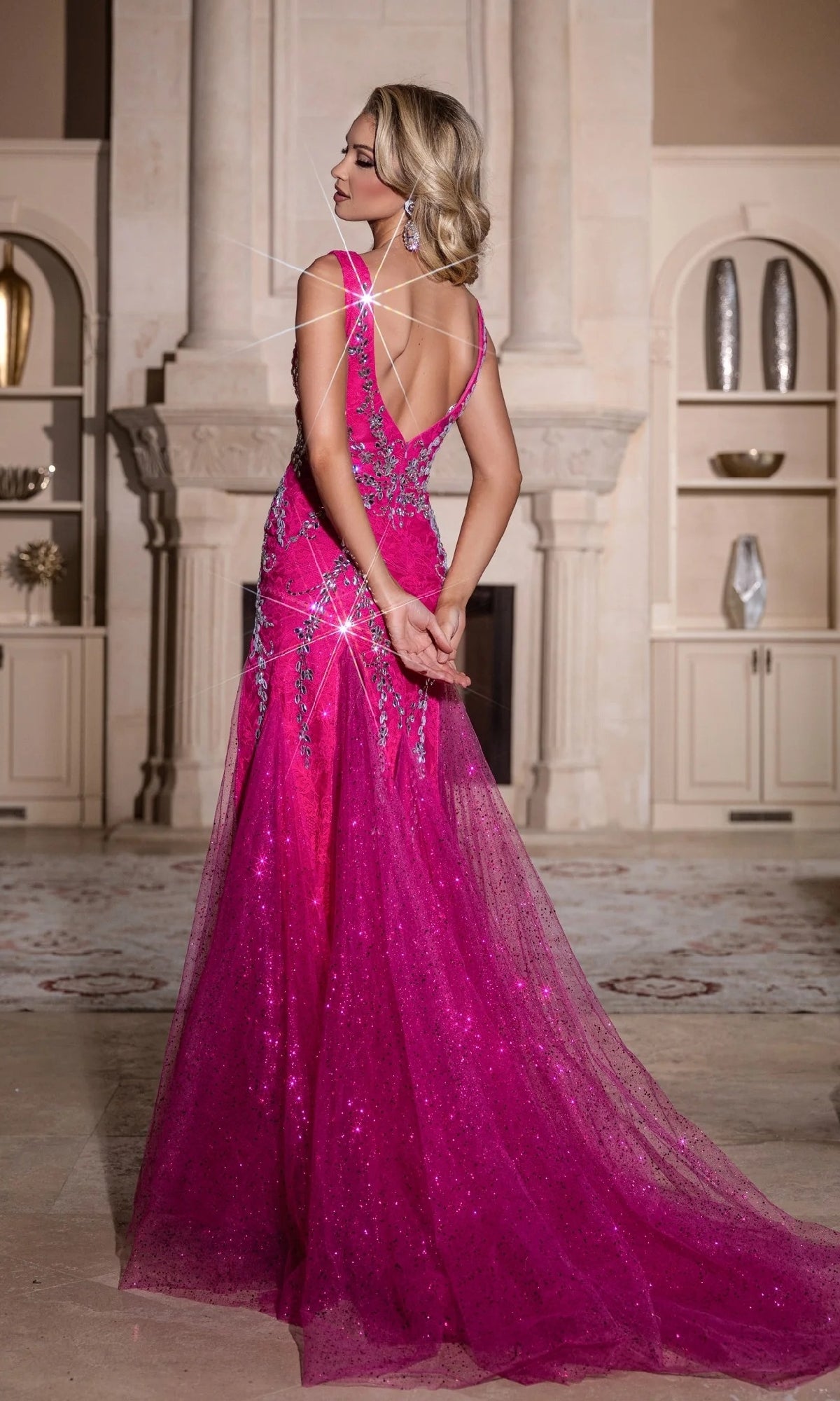 A woman in the Portia and Scarlett PS25289 long prom dress, a sparkly bright pink mermaid gown, stands with her back to the camera in an elegant room with shelves and decor.