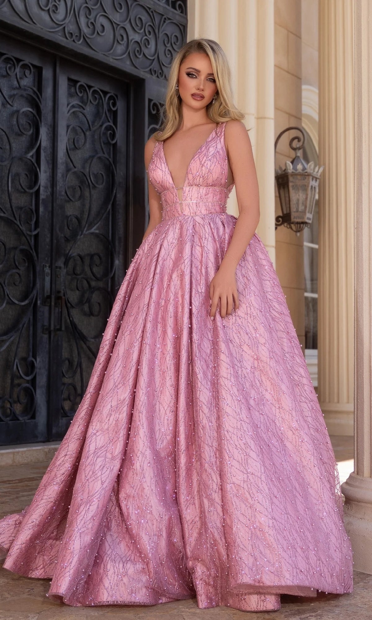 A woman stands before ornate doors wearing the Portia and Scarlett PS25316, a sleeveless, floor-length pink prom gown with a deep V-neckline and textured fabric.