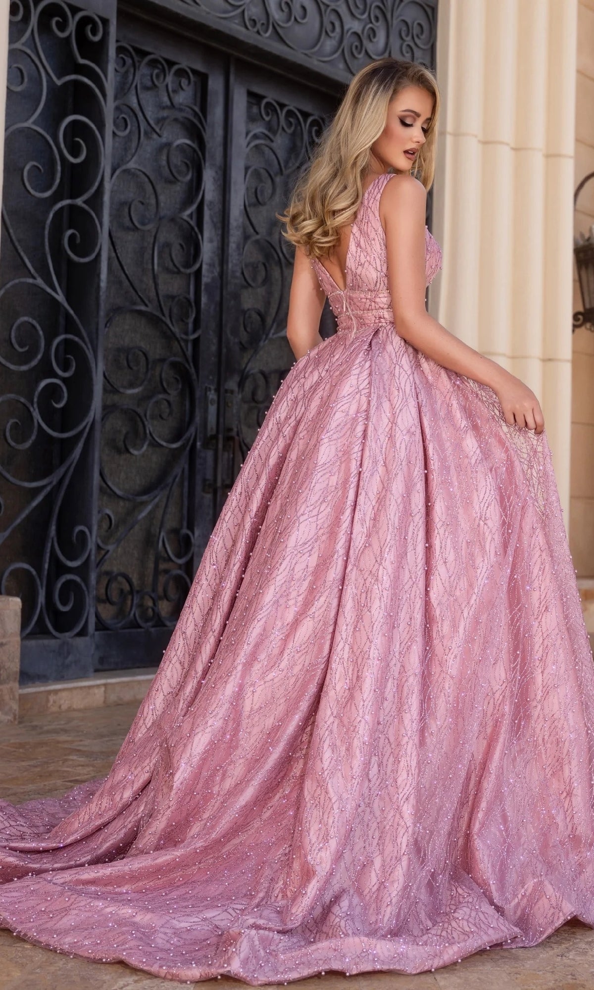 A woman wearing the Long Prom Dress: Portia and Scarlett PS25316, shimmering with delicate crystal beading, stands in front of ornate black doors, glancing over her shoulder.