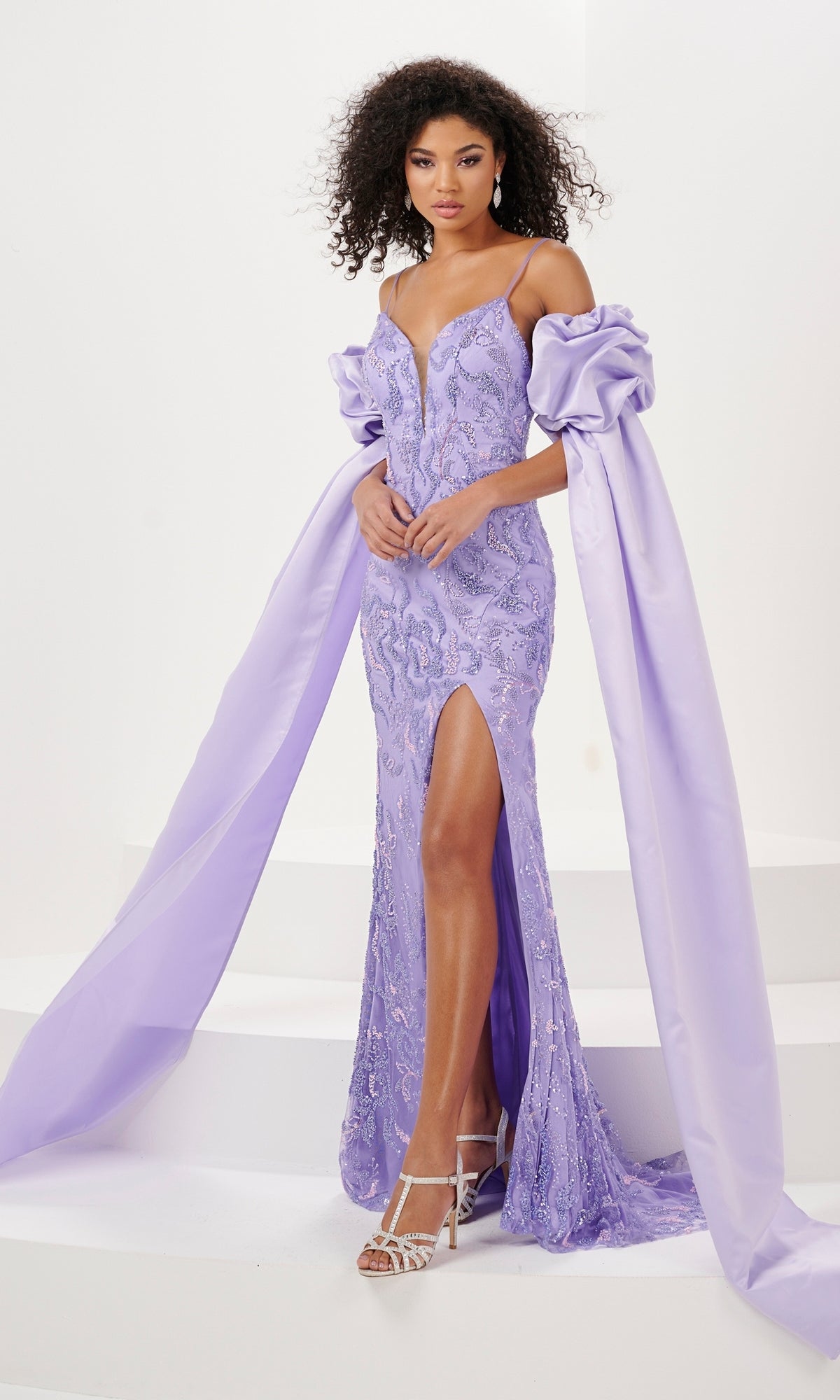 A woman wears the Sequin-Print Draped-Sleeve Long Prom Dress 14155—a lavender off-the-shoulder gown with puffed sleeves, a thigh-high slit, lace detailing, and long draped fabric trains for an elegant formal look.