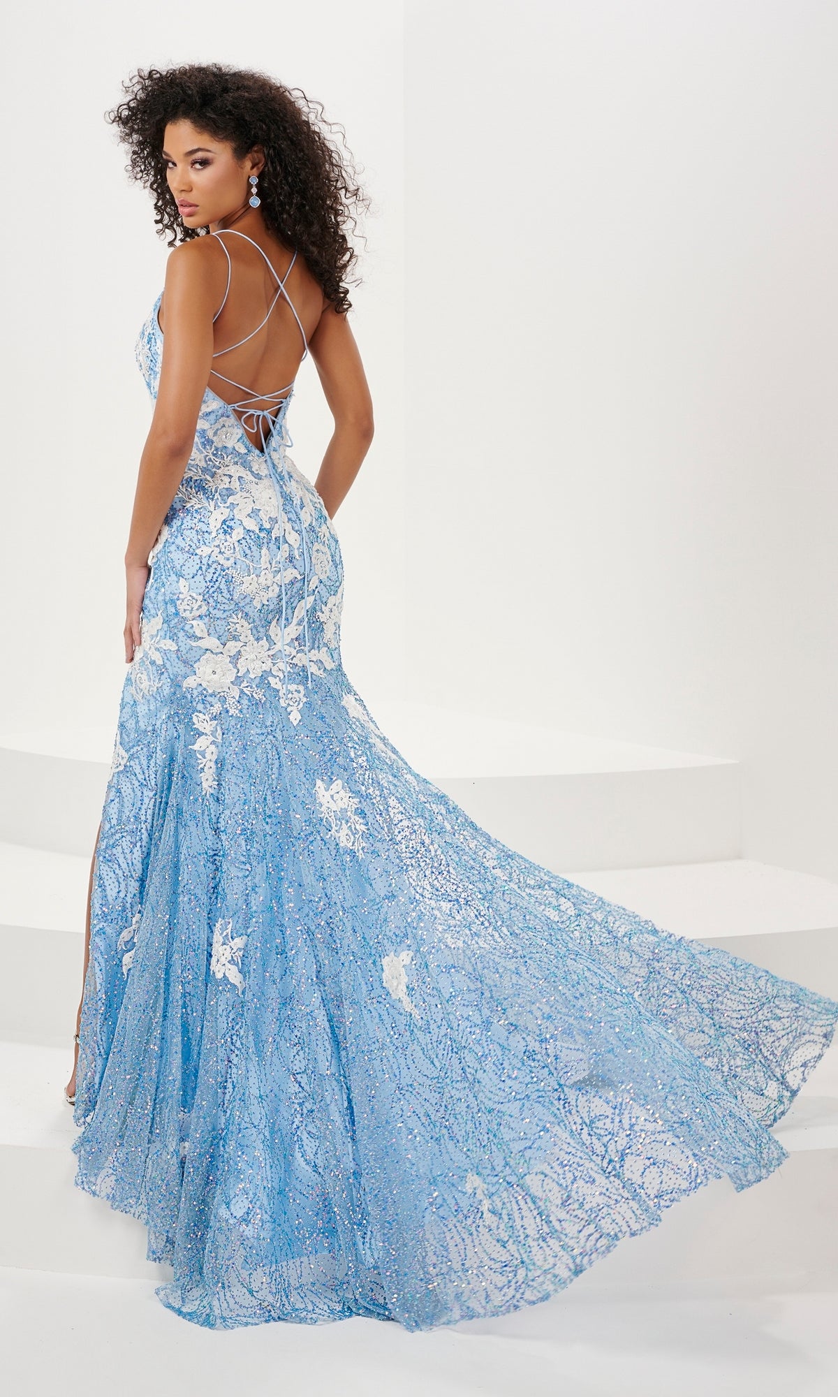 A woman models the Floral Cracked-Ice Sequin Long Prom Dress 14157, a light blue gown with floral patterns, a long train, and crisscross back straps, as she stands on white steps in a minimalist setting.