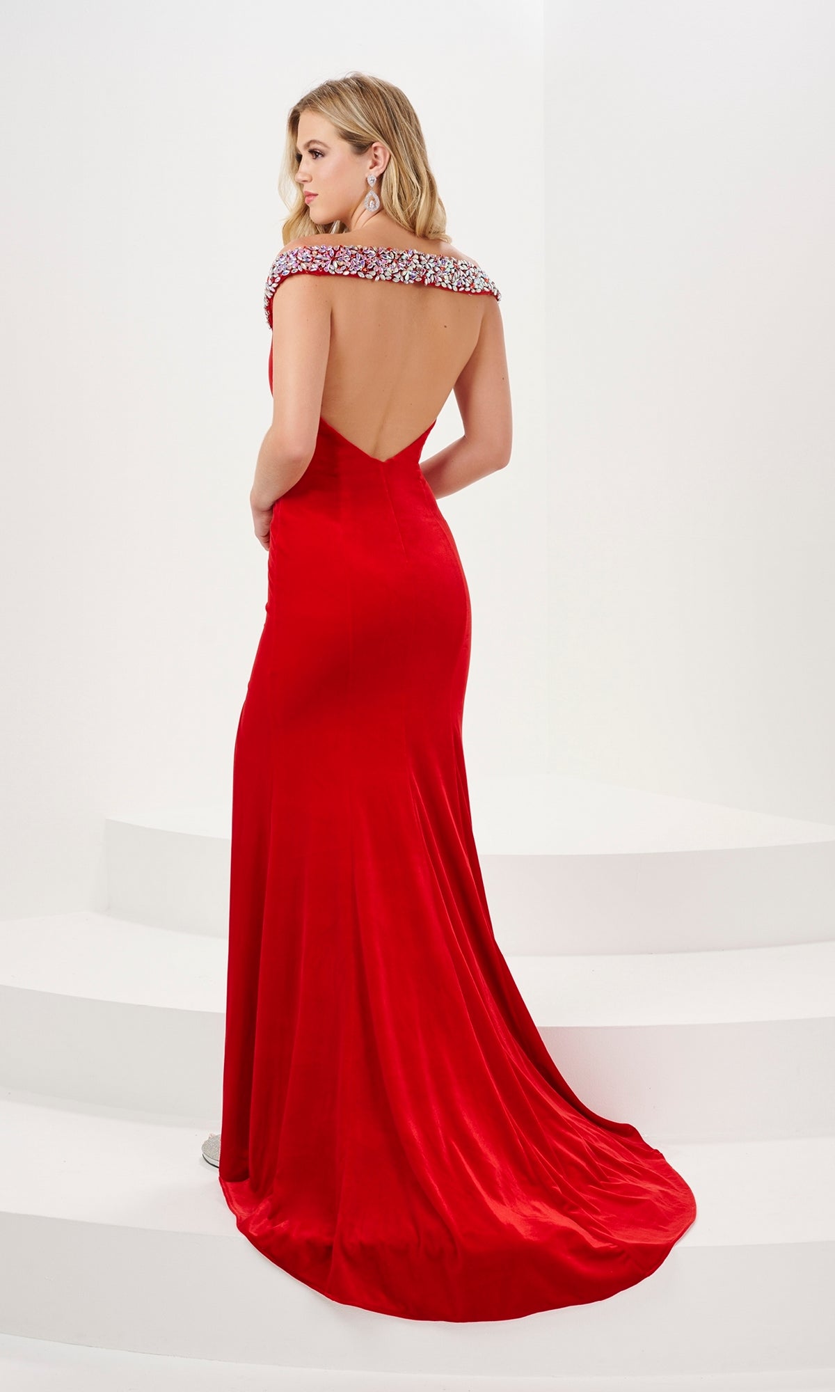 A woman stands on white steps, facing away in the Long Prom Dress 14195 by Panoply—a floor-length red stretch velvet gown with an open back and jeweled neckline.