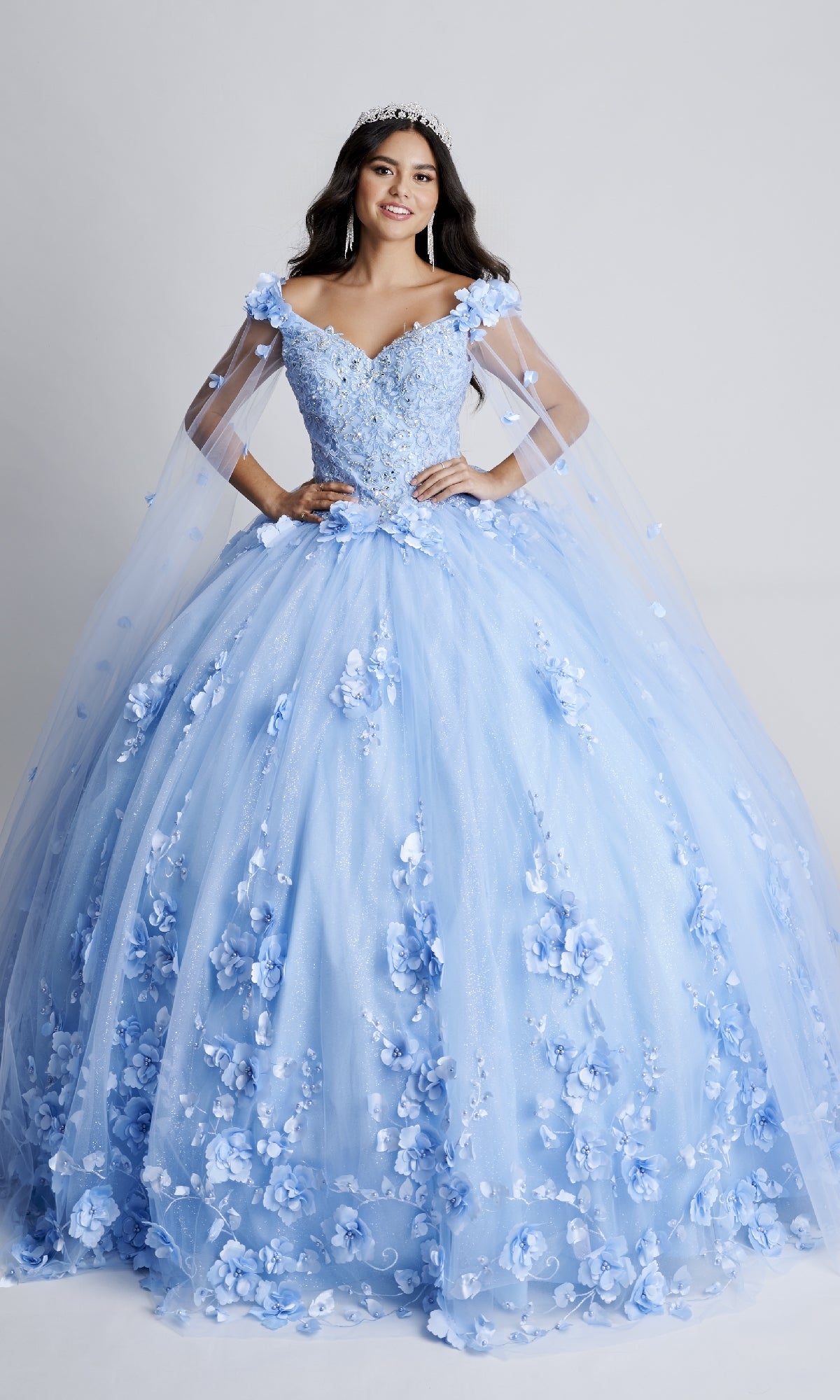 A young woman in the Nina Canacci Q100 long quinceanera dress—a light blue, off-the-shoulder ball gown with floral appliques and sheer cape sleeves—wears a tiara and stands against a plain background.
