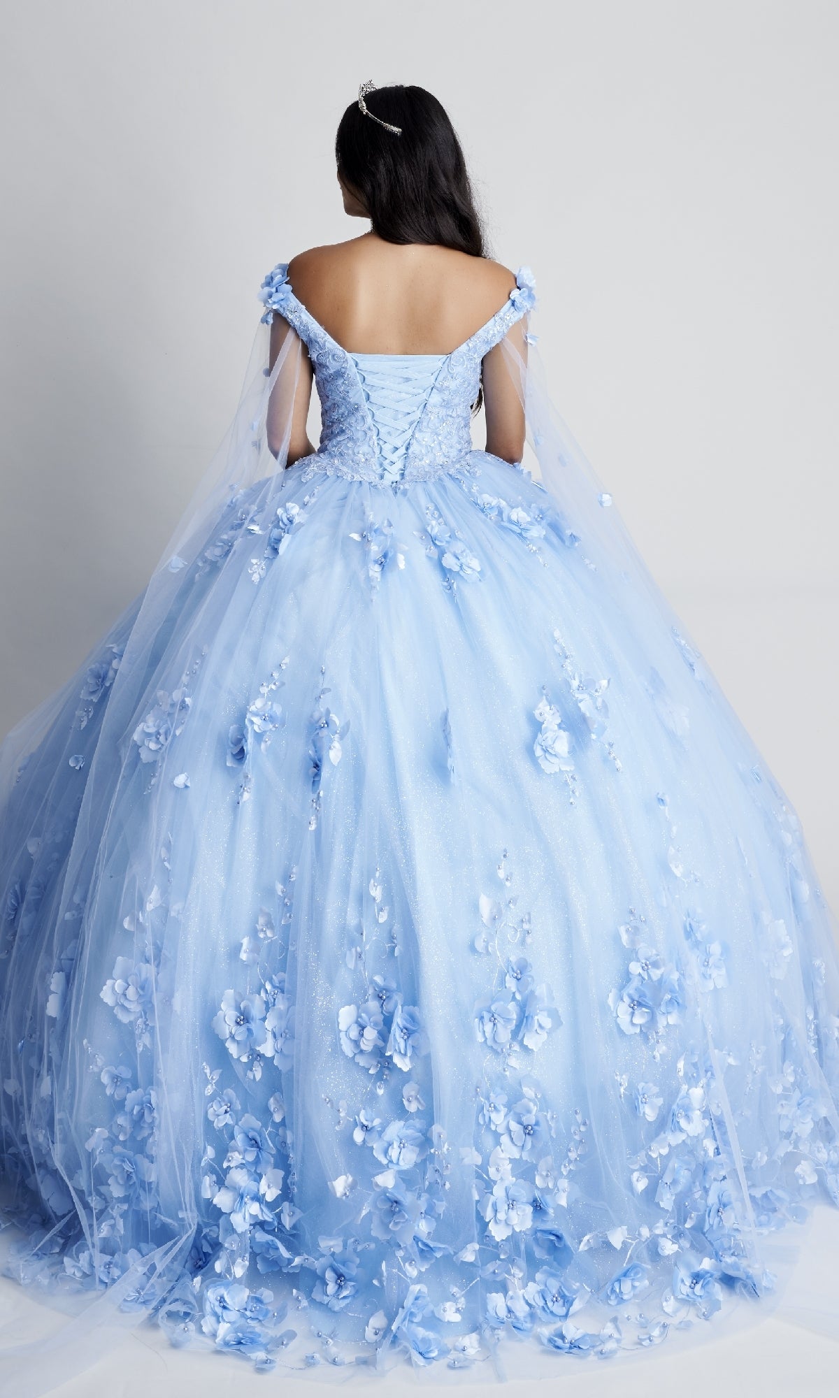 A person in the Nina Canacci Q100, a light blue off-the-shoulder quinceanera dress with floral lace appliqués, stands facing away against a plain background.