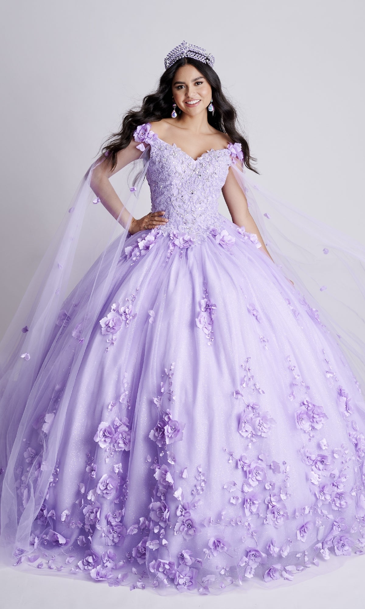 Young woman poses in the Nina Canacci Q100 long quinceanera dress, a light purple off-the-shoulder ball gown with floral lace, paired with a tiara and earrings, against a white background.